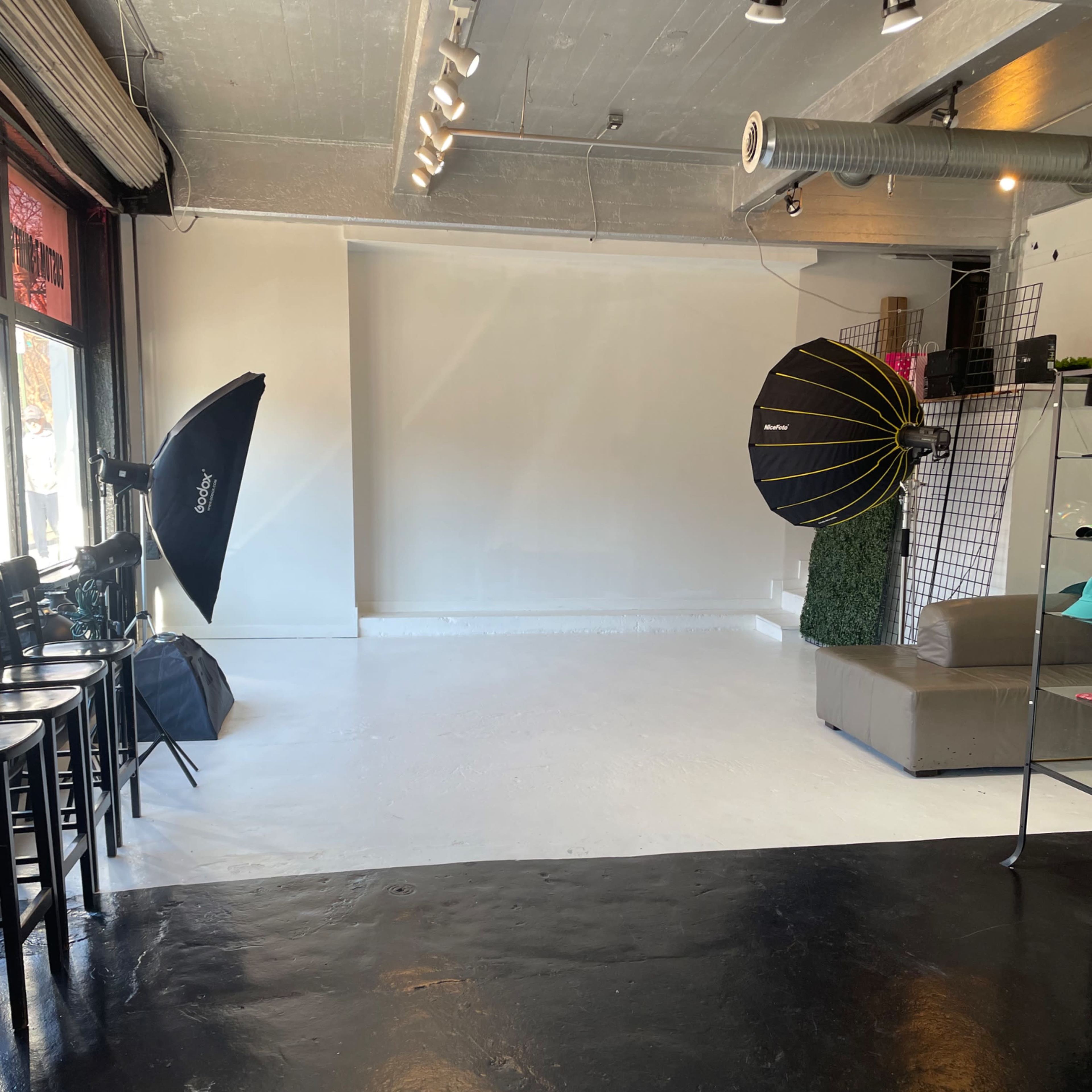 A spacious studio with a blank white backdrop, two softbox lights, and a seating area featuring a sofa and chairs.