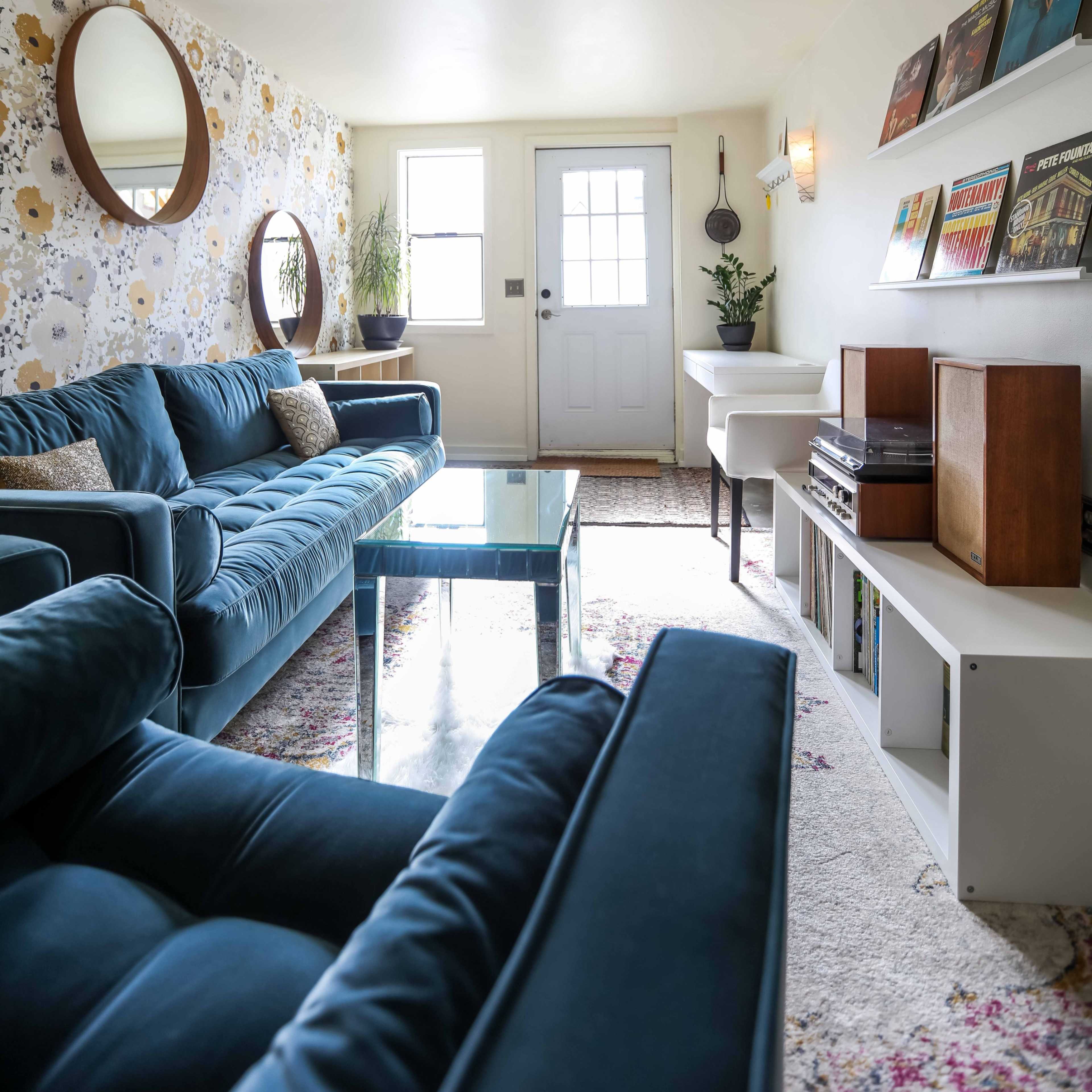 A living room with blue sofas, a glass coffee table, a door leading outside, and shelves displaying records and books.