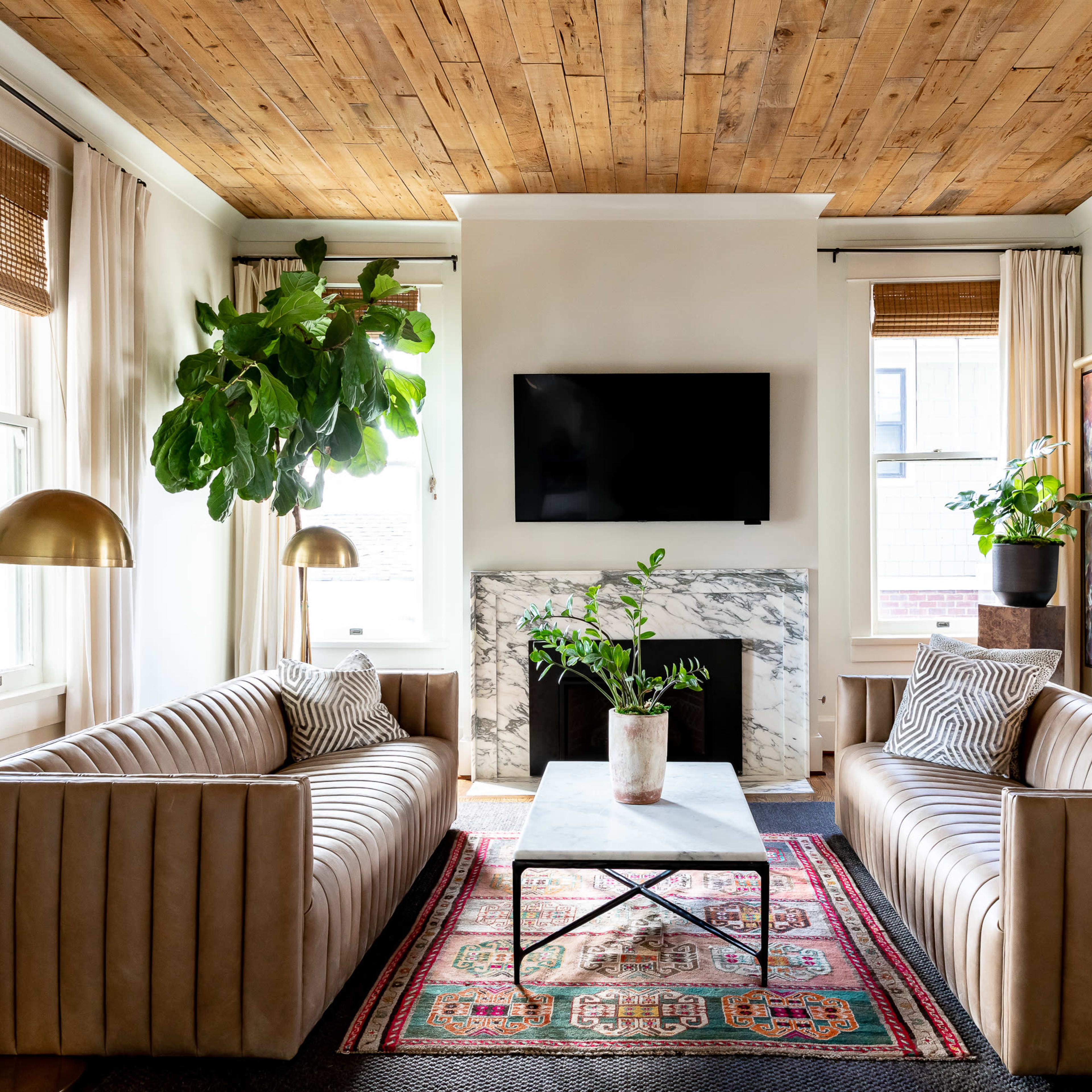 A cozy living room featuring two beige sofas, a coffee table with a plant, a marble fireplace, and a mounted television on a wooden ceiling backdrop.