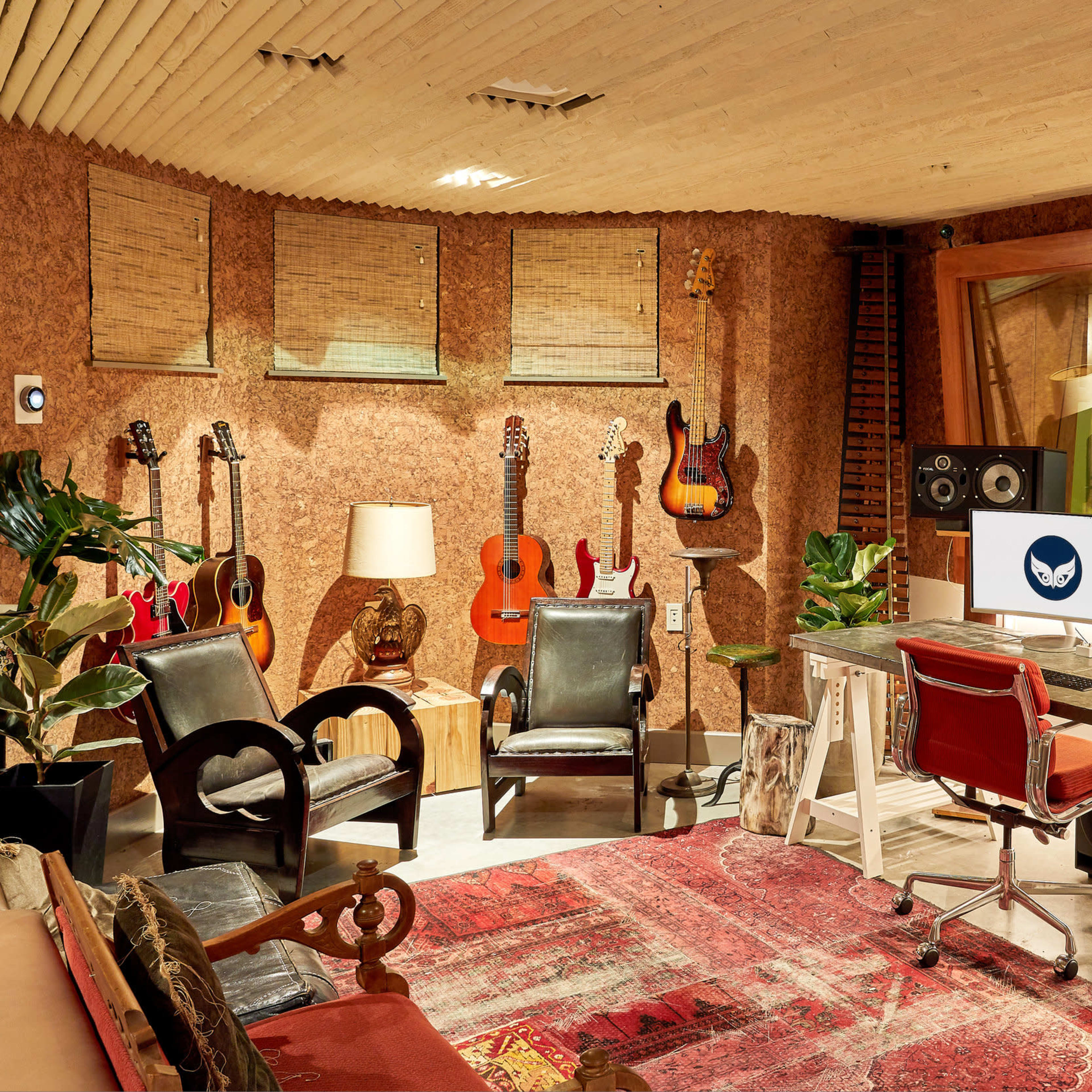 A music studio with a desk featuring dual monitors, audio equipment, and various guitars displayed on the walls, surrounded by seating and decorative elements.