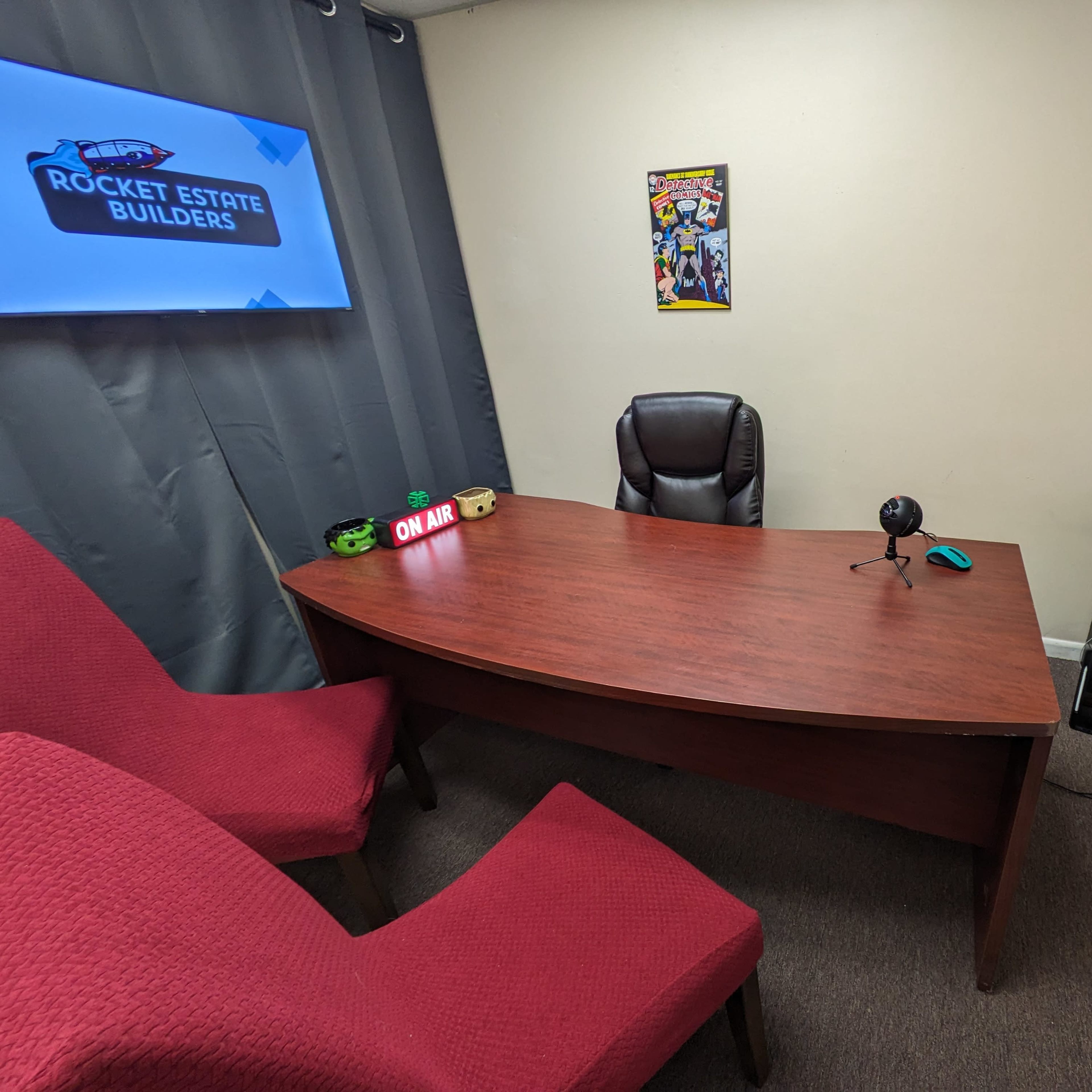 A small office space featuring a wooden desk with a microphone, a laptop, and two red chairs, set against a gray curtain and a wall-mounted screen displaying a logo.