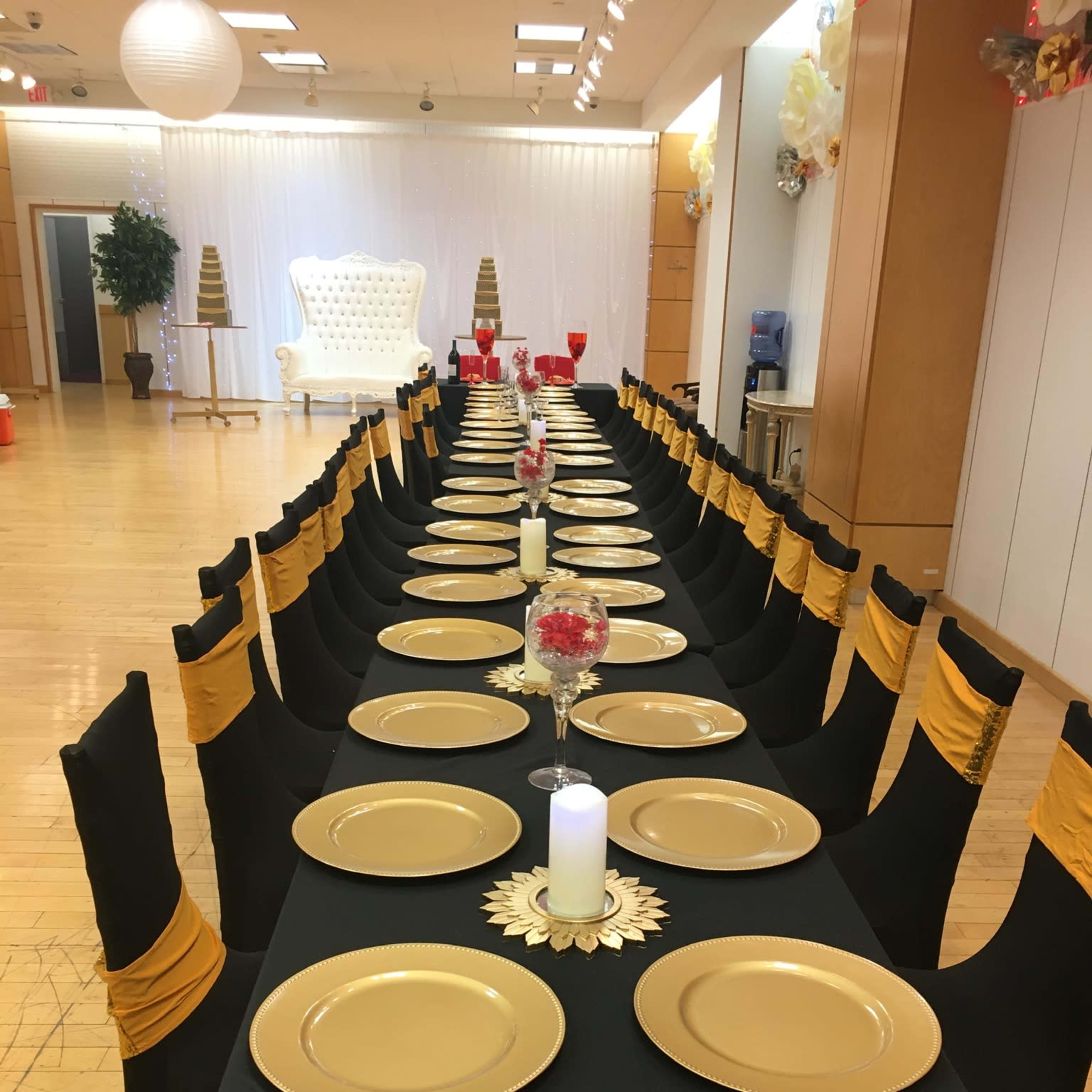 A long dining table is set with gold plates and candles, surrounded by black chairs with yellow sashes in a decorated event space.
