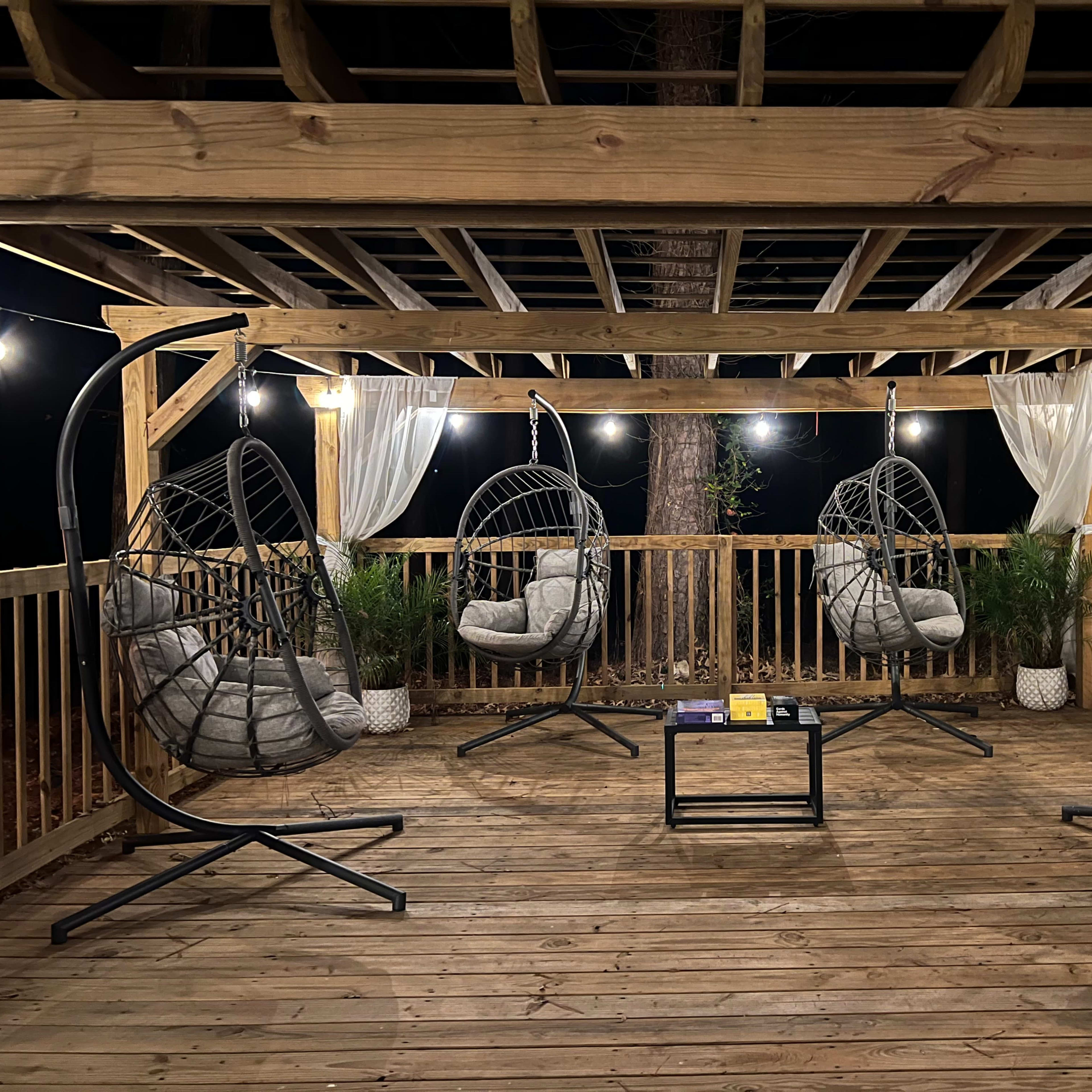 A wooden deck featuring four hanging chairs, a small table, and string lights illuminating the area at night.