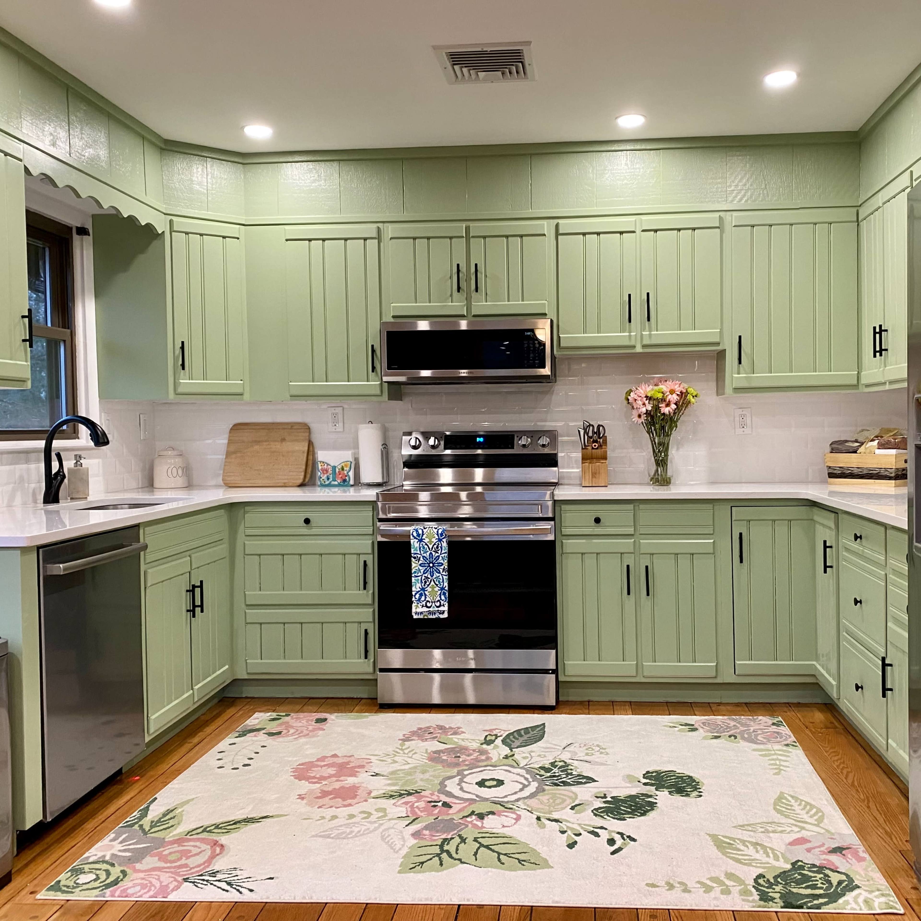 A well-organized kitchen features green cabinetry, a stainless steel stove, and a floral rug on the wooden floor.