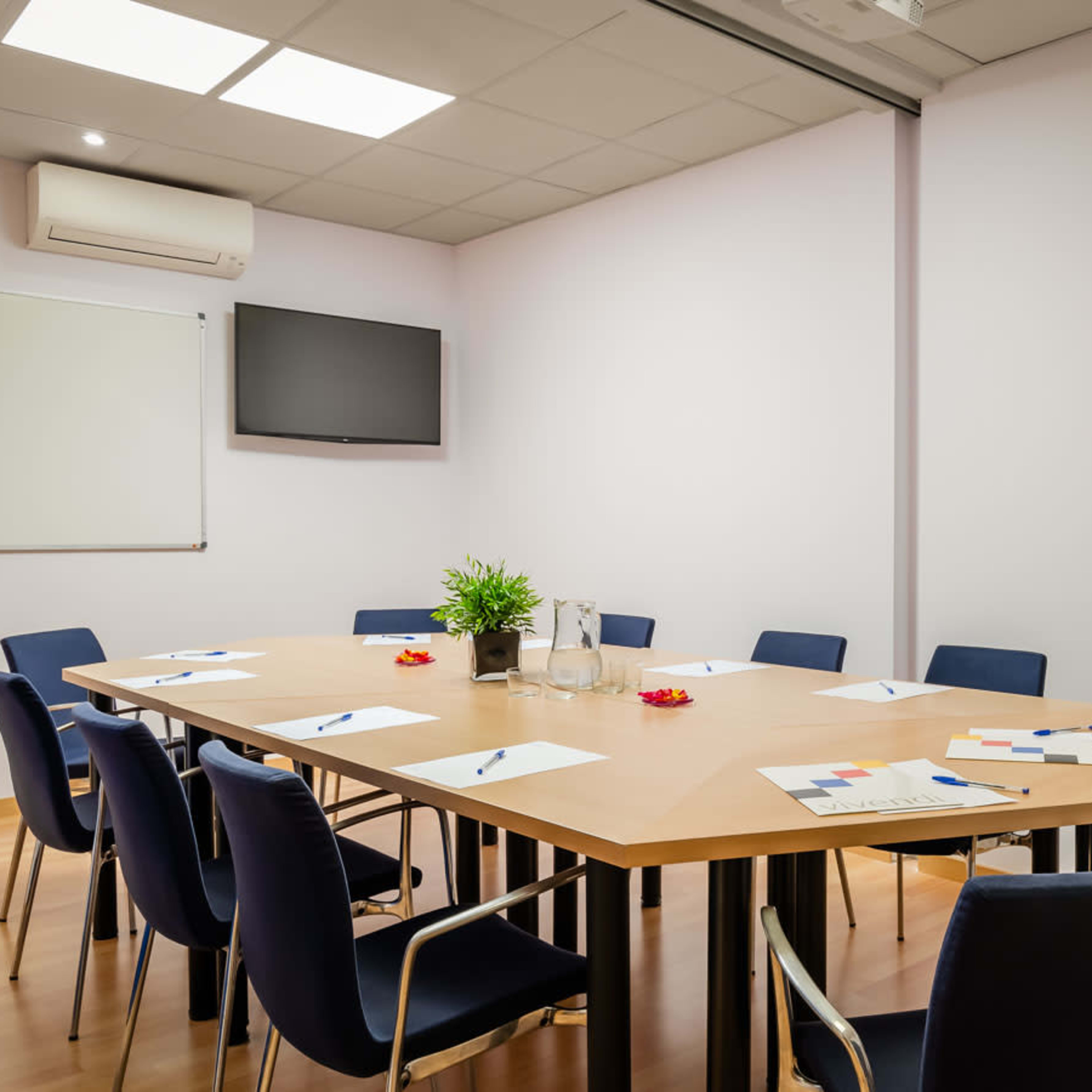 A conference room features a large table surrounded by eight chairs, with a television mounted on the wall and a whiteboard in the corner.