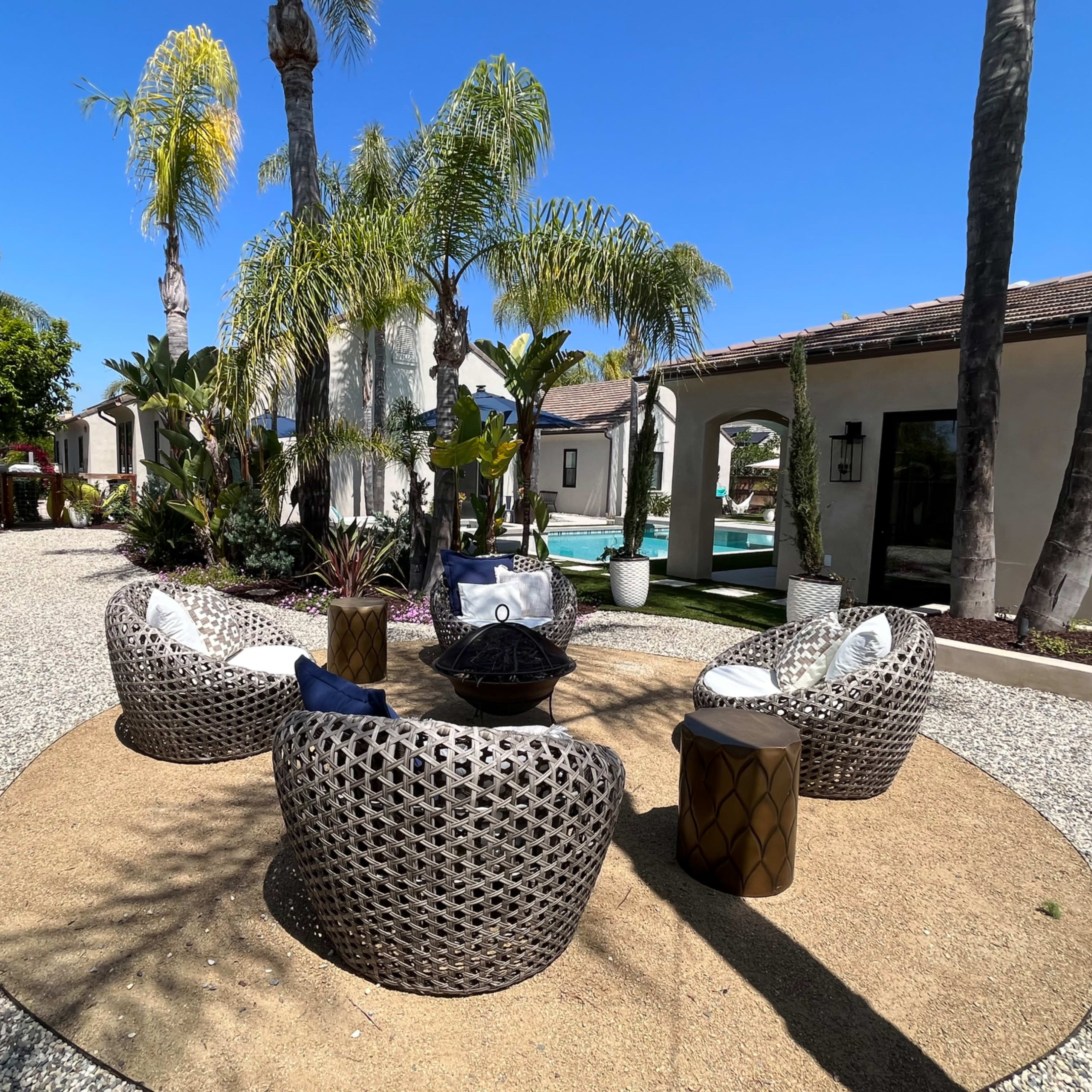 A circular seating area with four woven chairs and a central fire pit is surrounded by palm trees and landscaped greenery.