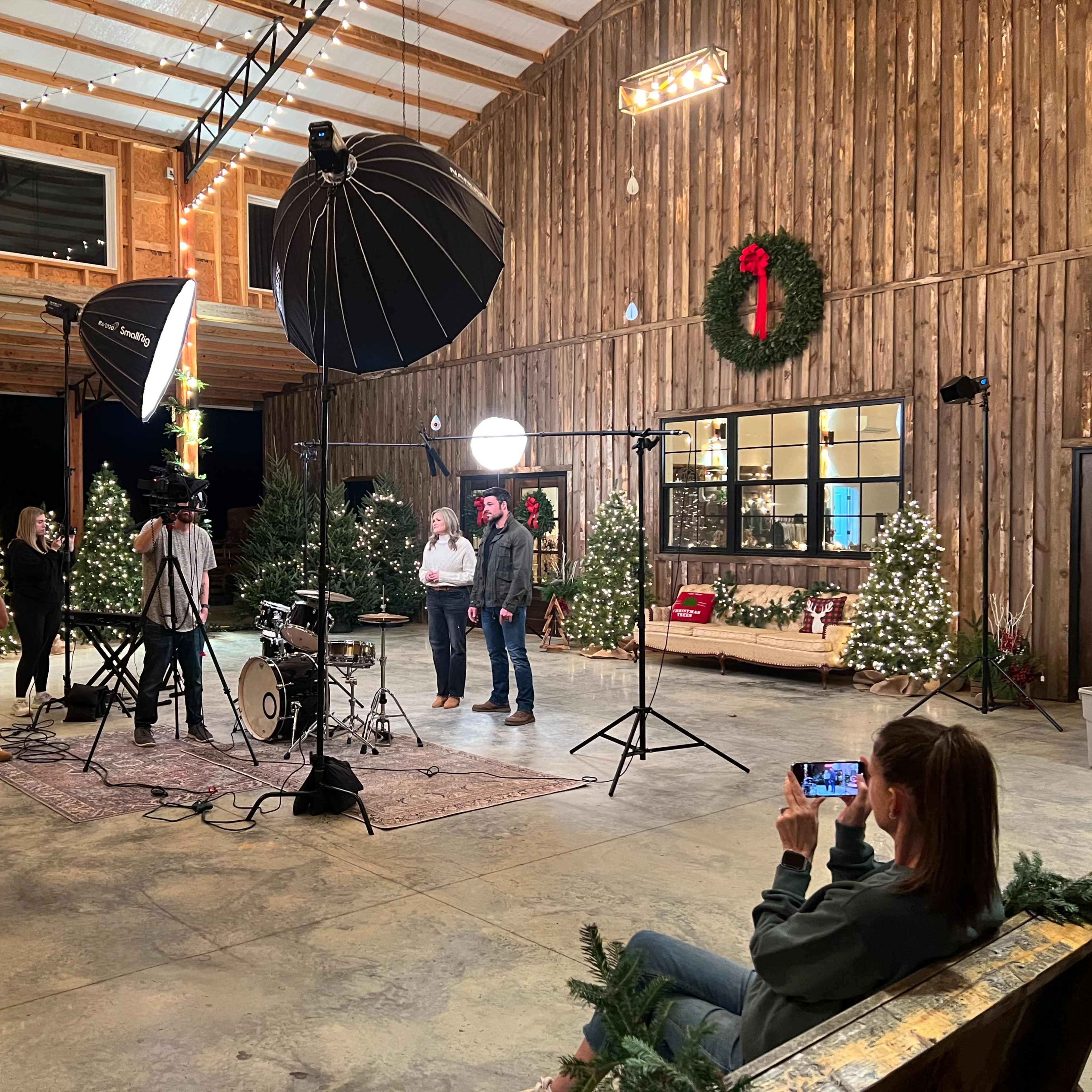 A group of people gathers around a photography setup inside a decorated barn with Christmas decorations and studio lighting equipment.