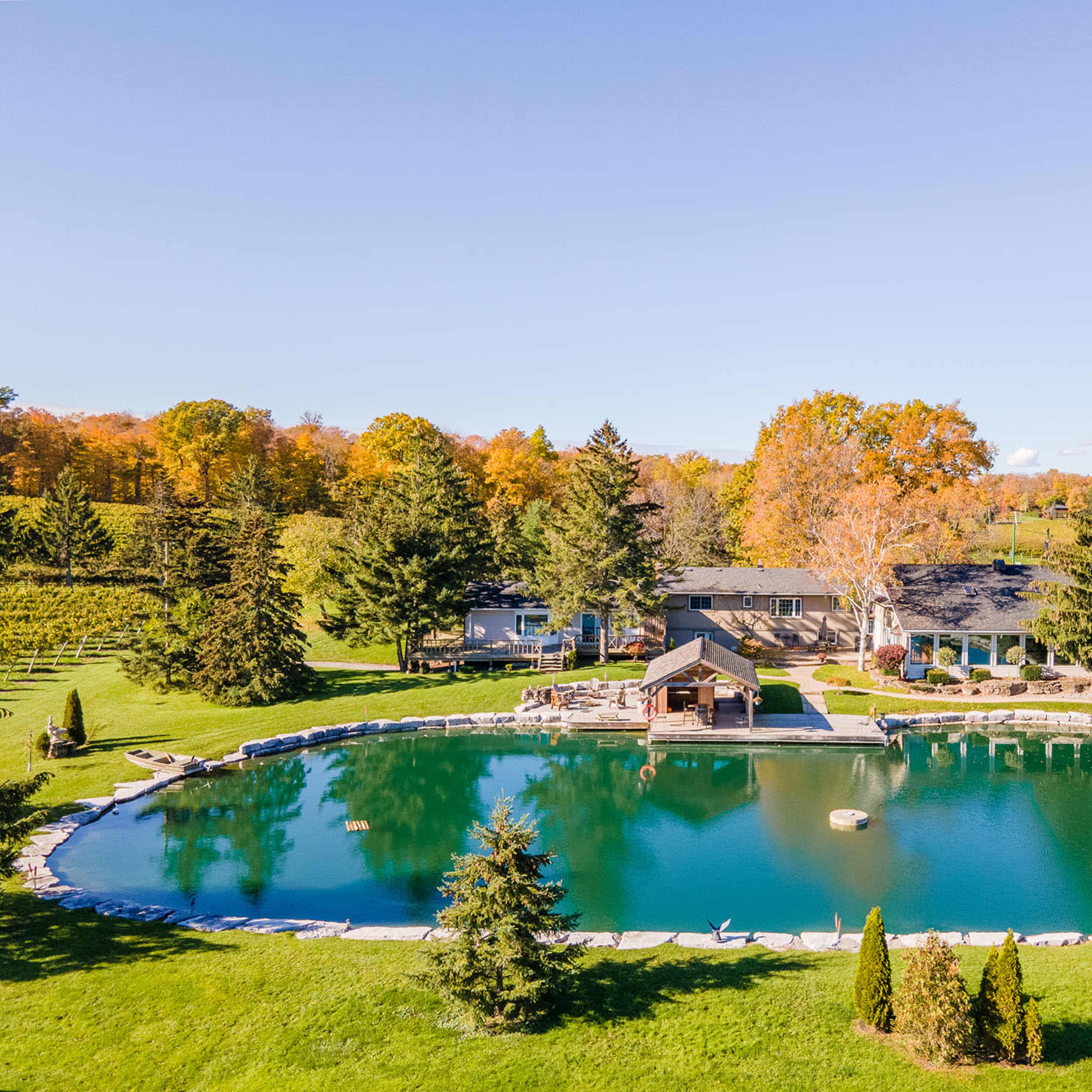 A large, serene pond surrounded by trees and a stone edge is located in front of a house and a gazebo.