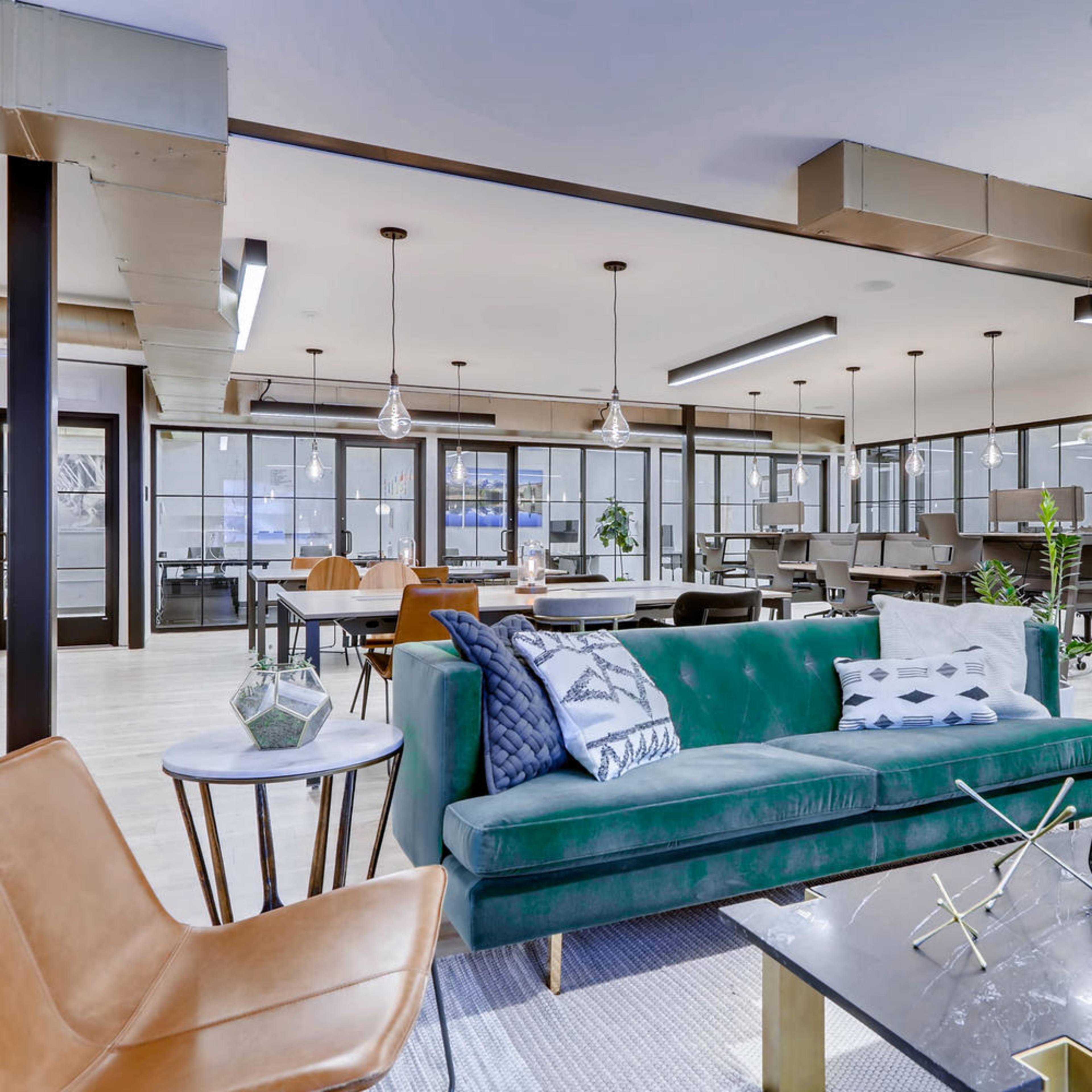 A modern office lounge area with a green sofa, brown chairs, and various tables arranged in a bright space featuring large windows.