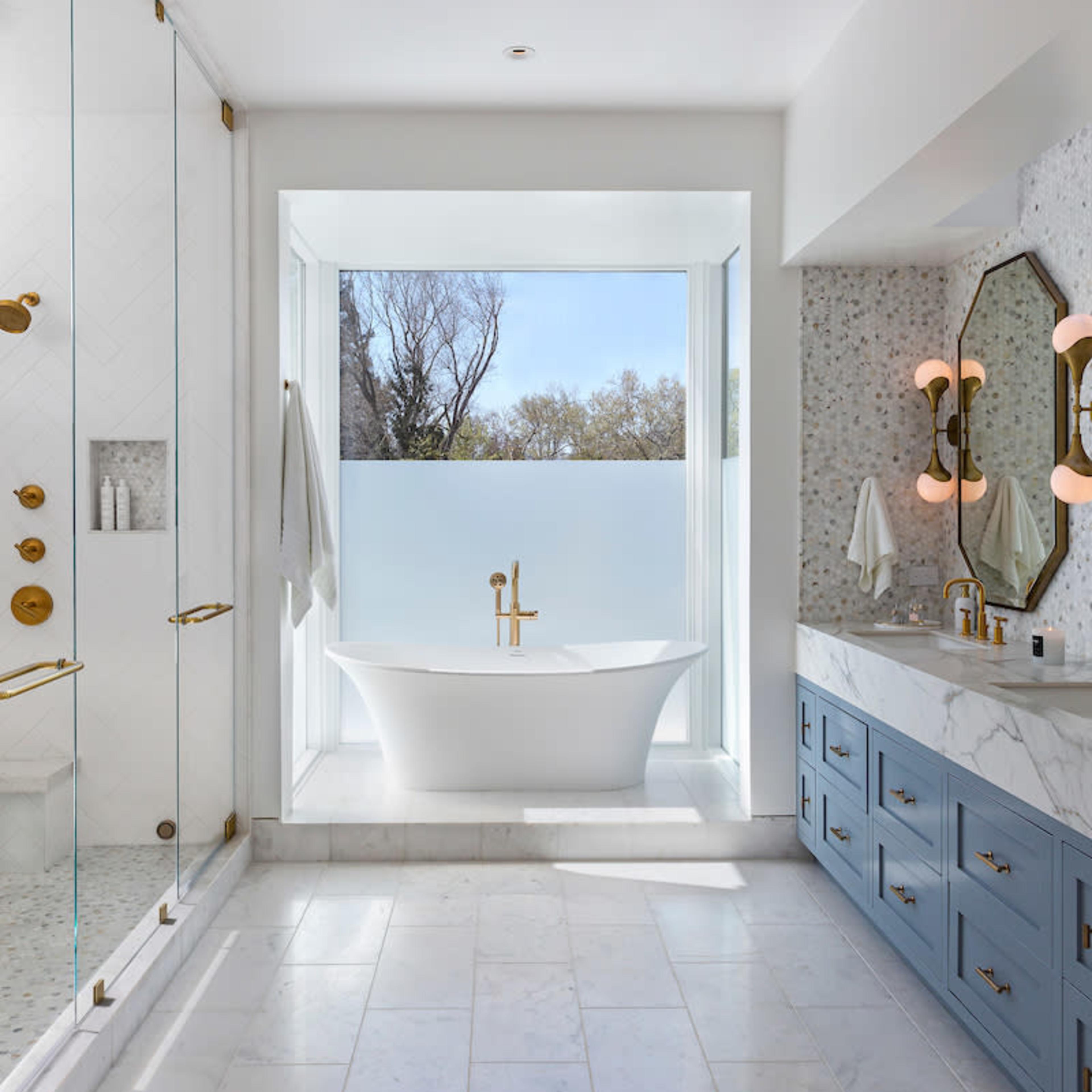 The bathroom features a freestanding bathtub next to a large window, a glass shower enclosure, and a dual-sink vanity with blue cabinets.