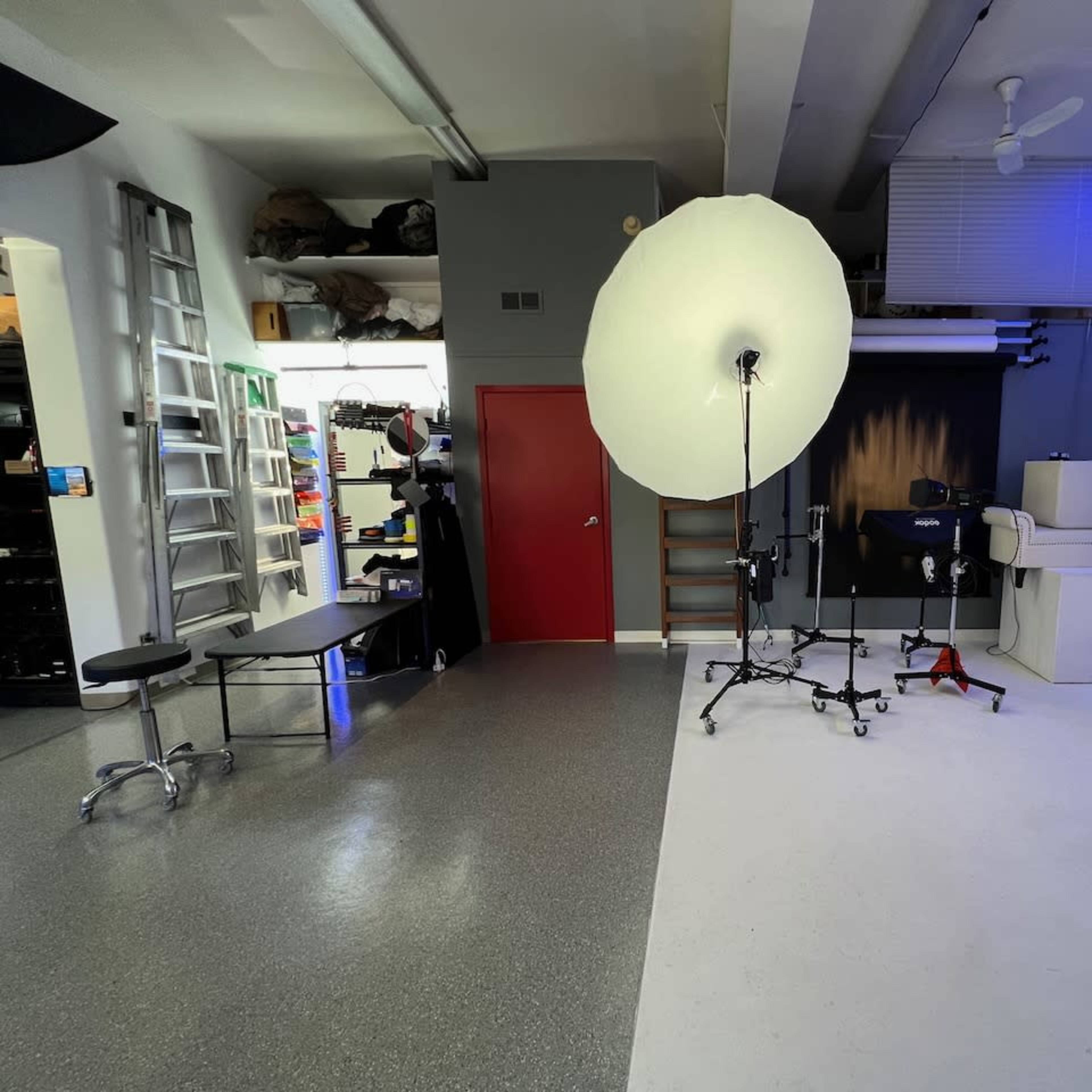 A photography studio with a large light umbrella on a stand, a white backdrop, and various equipment placed in the corners.