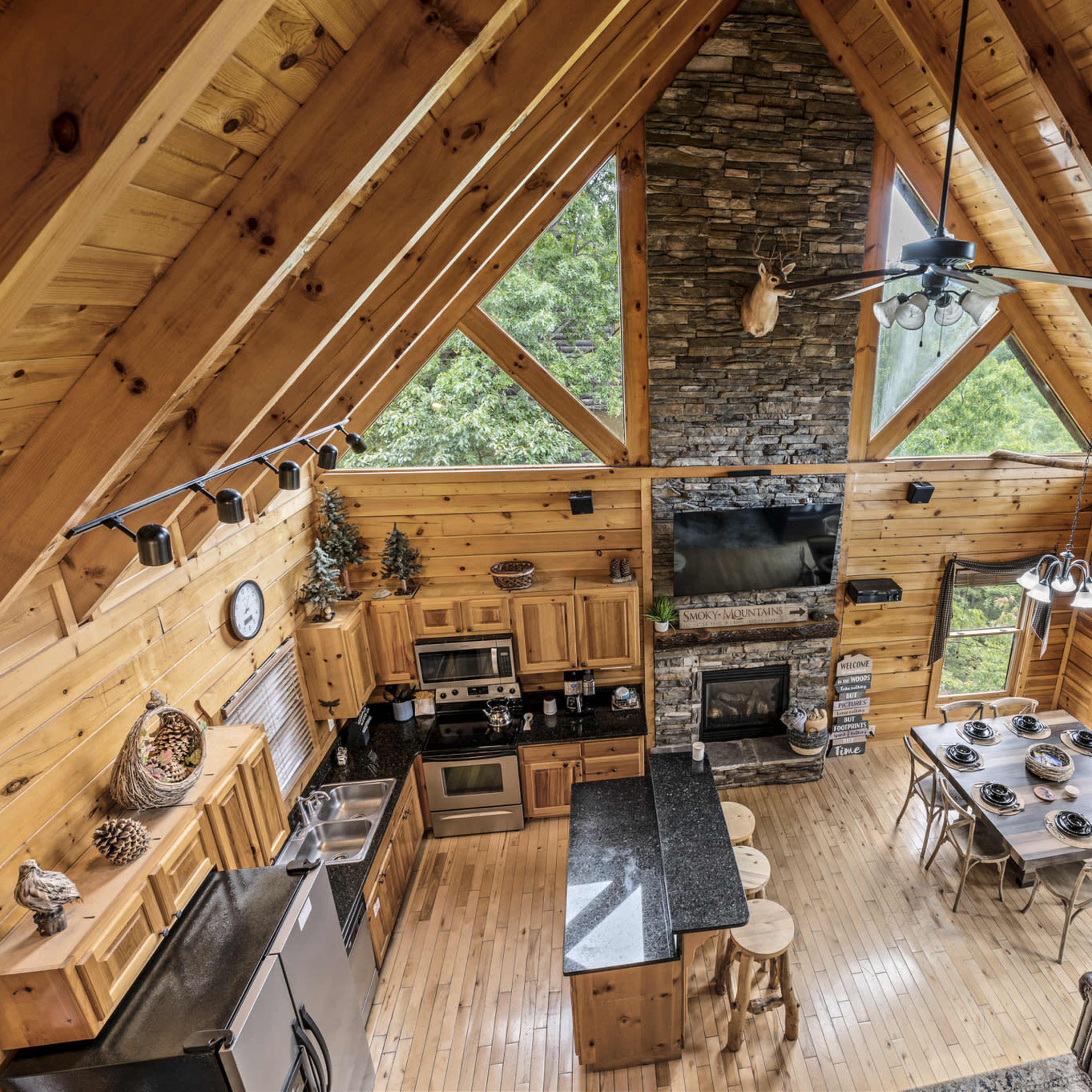 The interior of a spacious wooden cabin features a kitchen with modern appliances, a large dining table, and a stone fireplace under a high, vaulted ceiling.