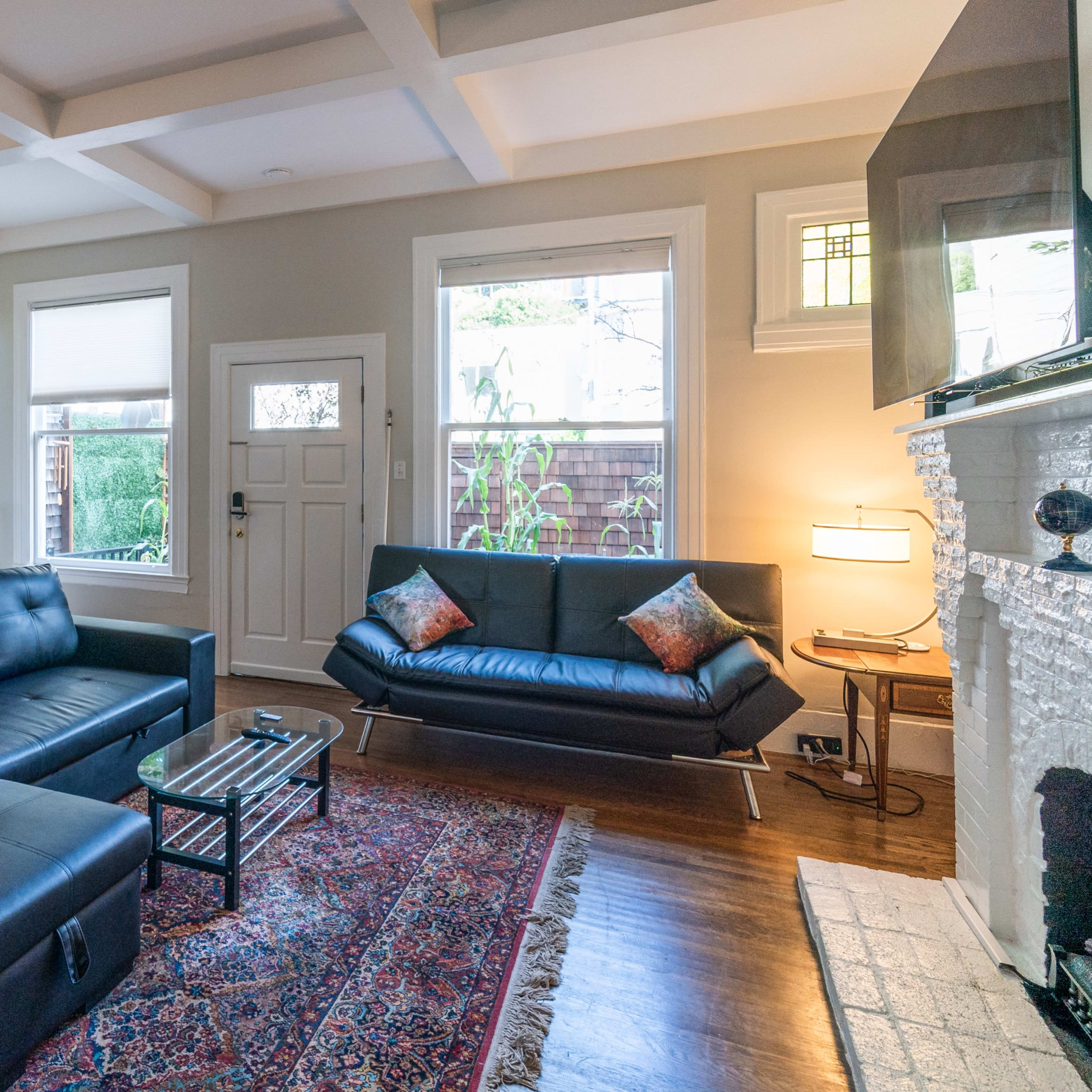 The living room features a black leather sectional sofa, a glass coffee table, and a light-colored stone fireplace, illuminated by natural light from the windows.