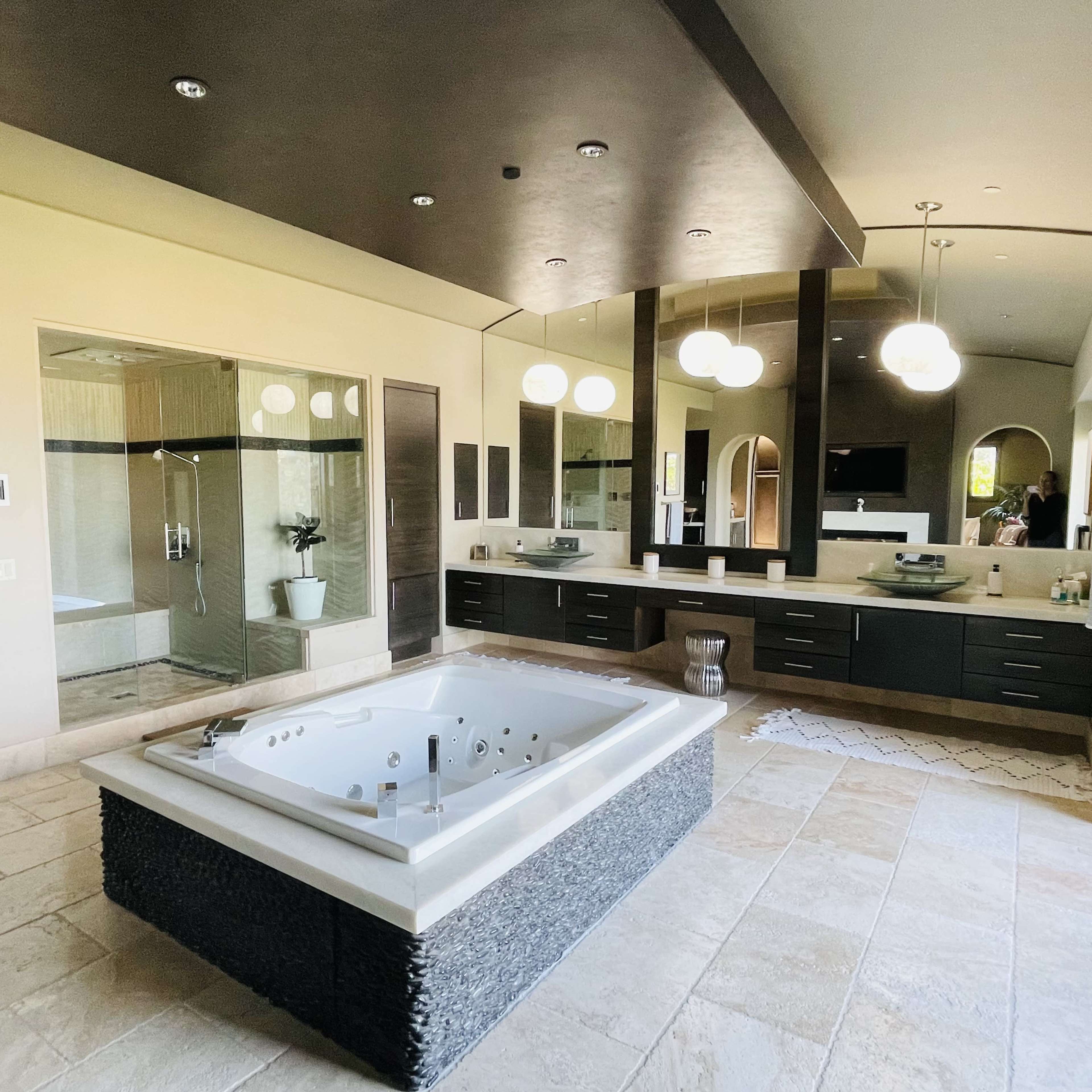 A spacious modern bathroom featuring a large jetted bathtub, a walk-in shower, and dual sinks surrounded by dark cabinetry and bright lighting.