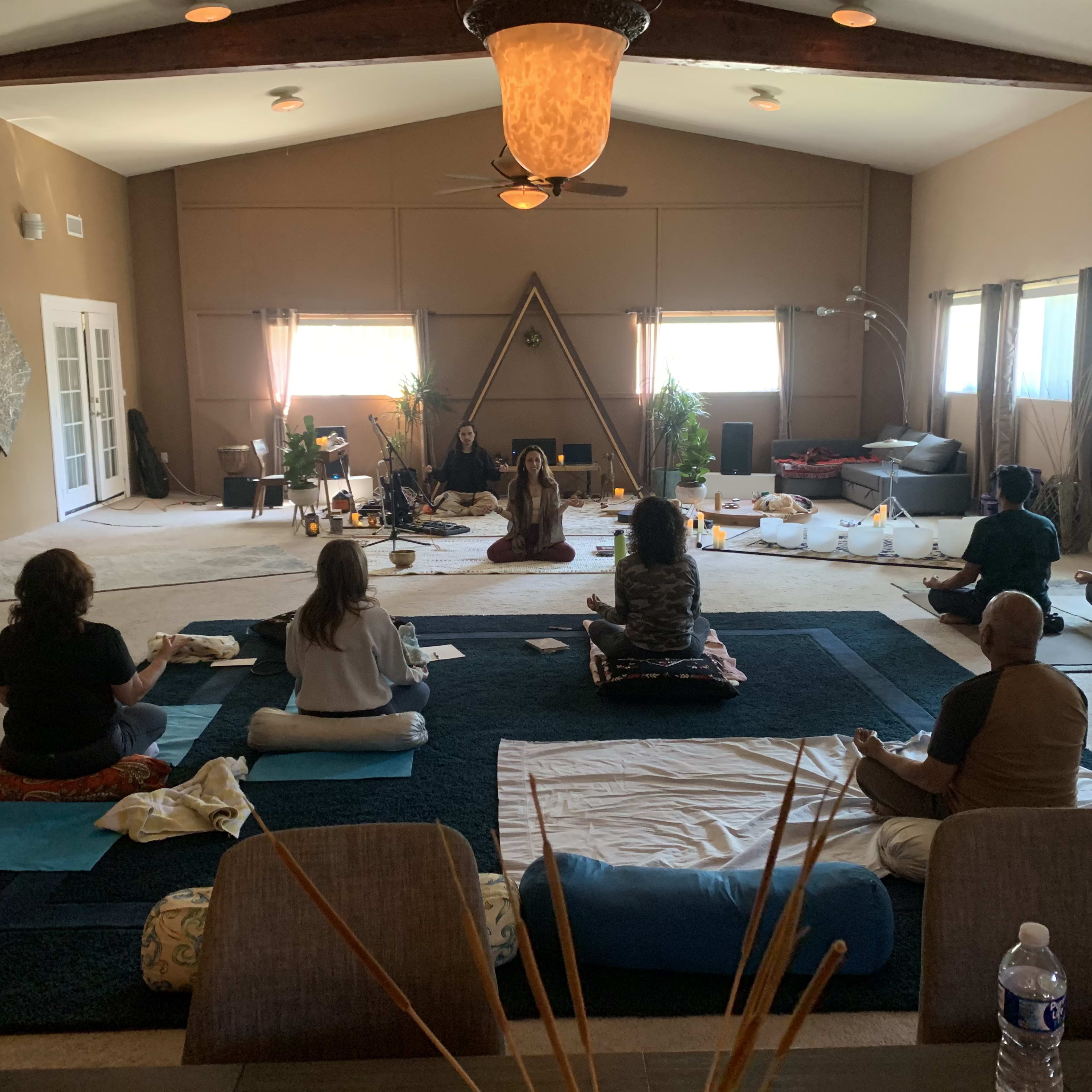 A group of individuals sits on mats in a serene, spacious room arranged for a meditation or wellness session.
