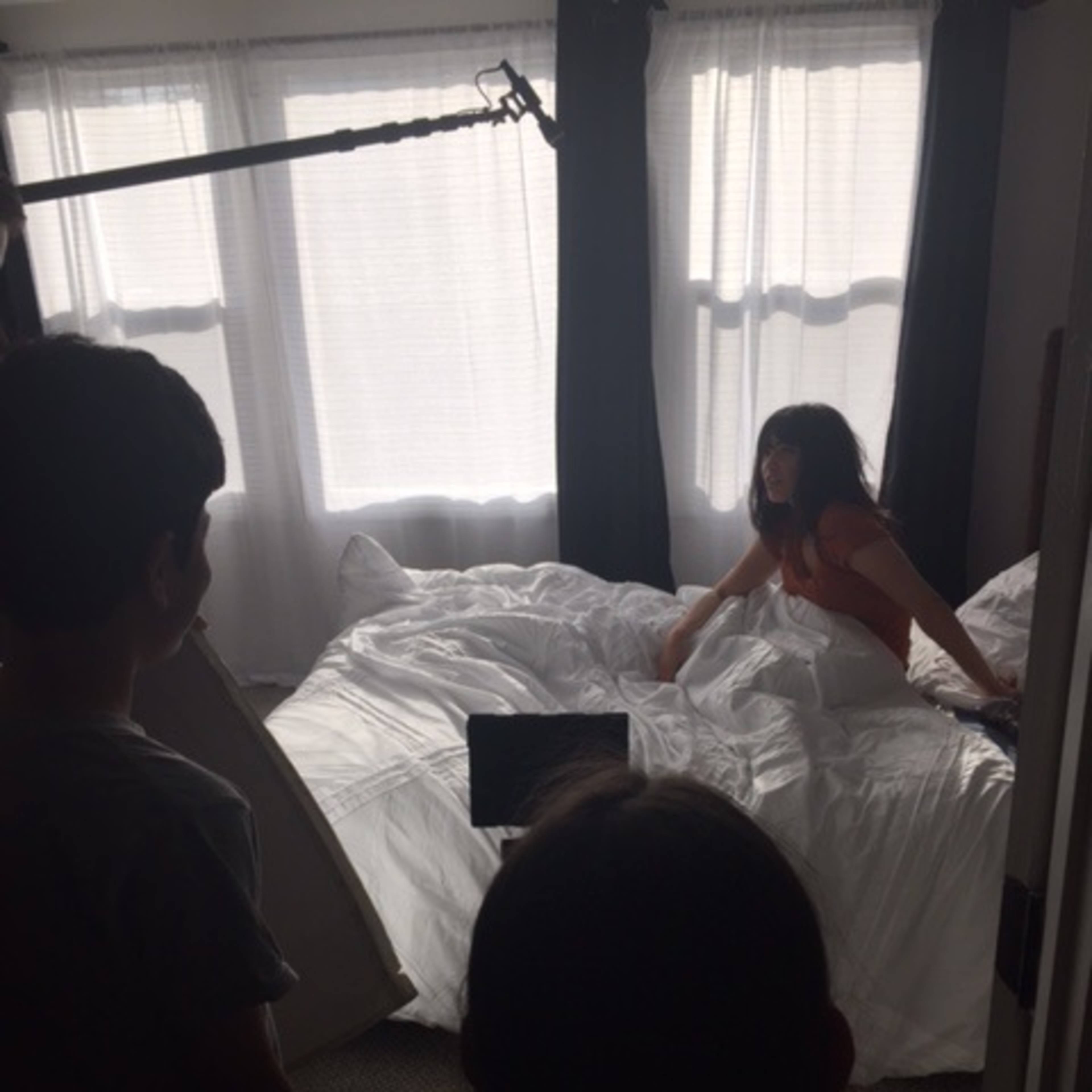 A woman sits on a bed covered with white bedding while two people hold equipment nearby.