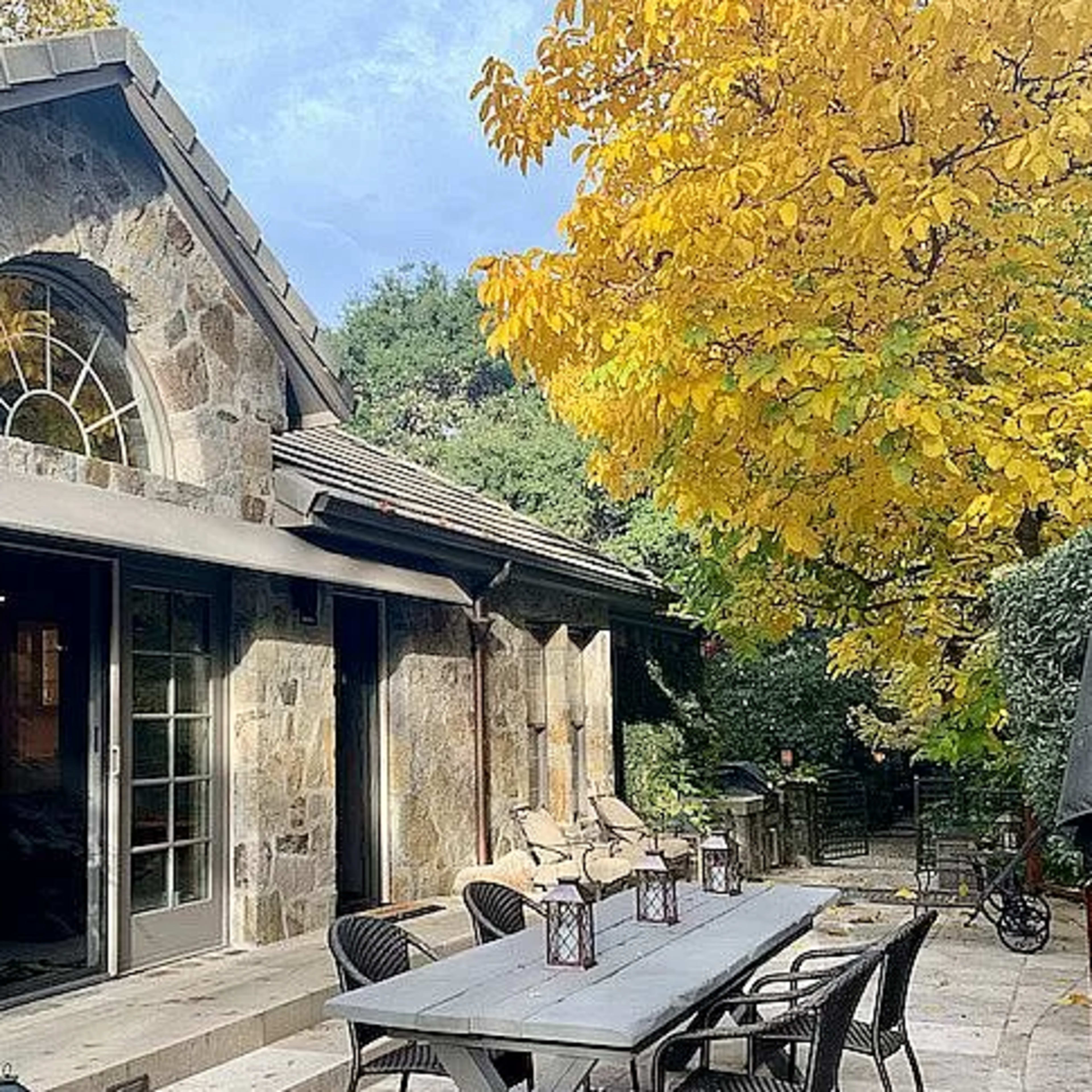 A stone house with large windows, a shaded outdoor dining area featuring a long table and chairs, and a vibrant yellow tree nearby.