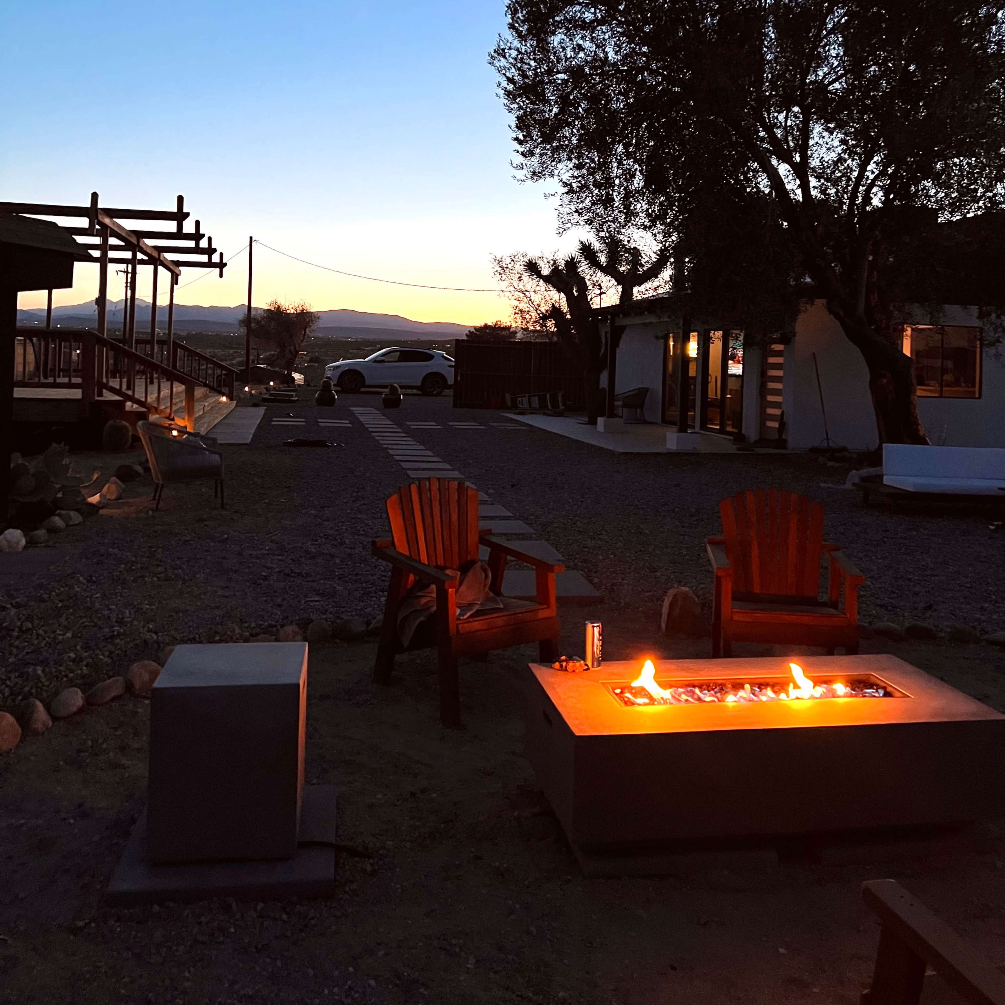 A gravel yard with a fire pit and Adirondack chairs, framed by a tree and a house against a twilight sky.