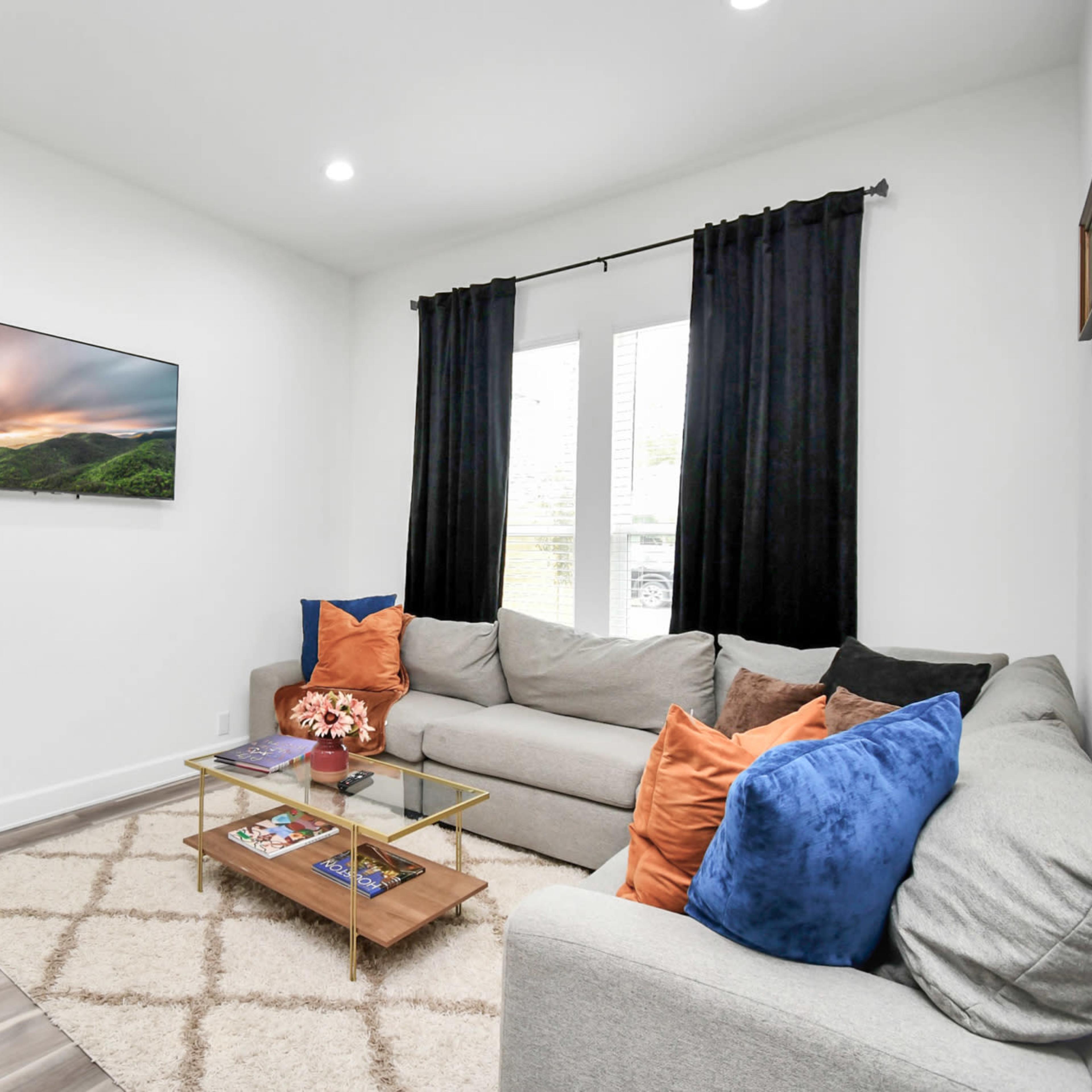 The living room features a gray sectional sofa, a glass coffee table with magazines, a large mirror, and a wall-mounted television above a decorative stone feature.