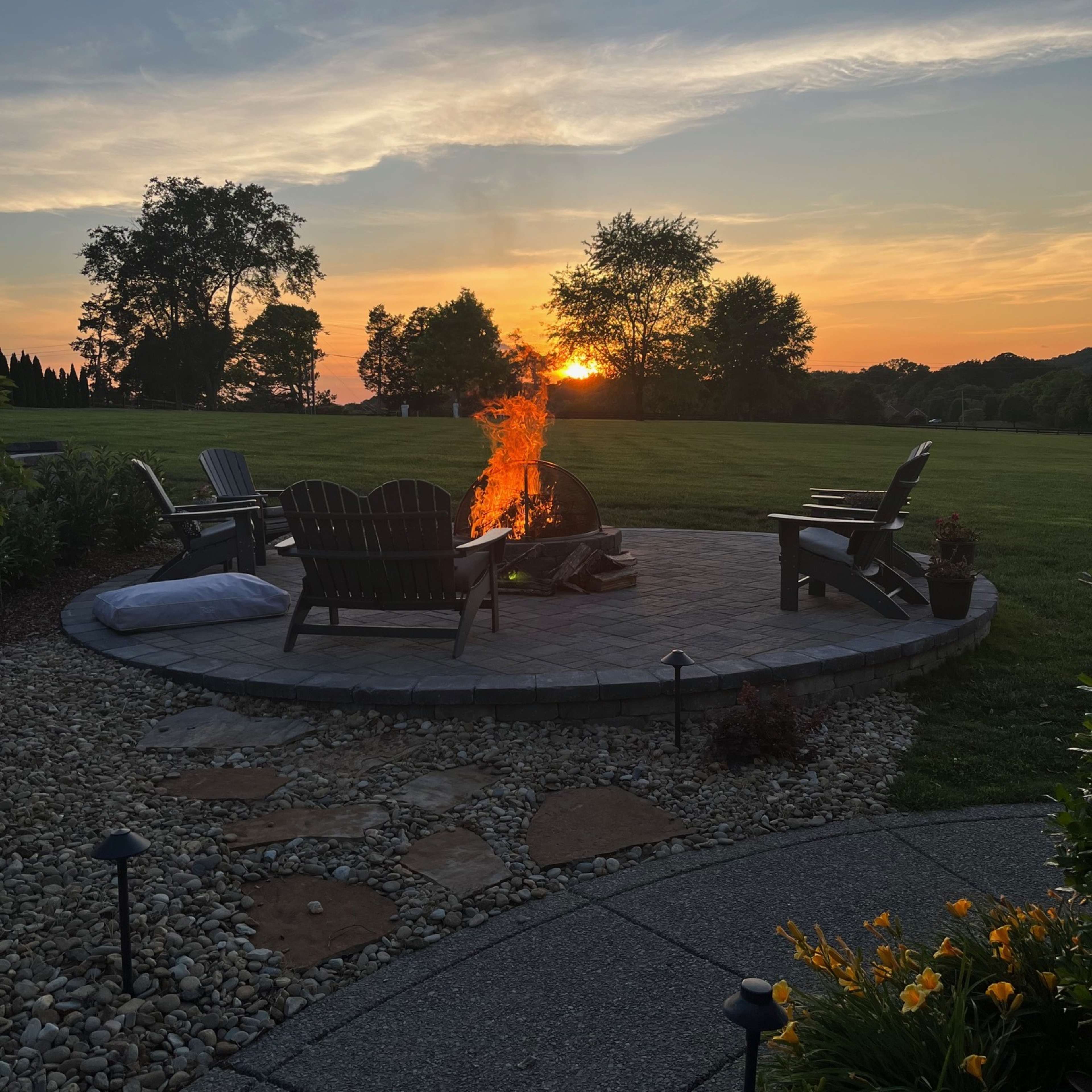 A fire burns in a stone fire pit surrounded by wooden chairs as the sun sets in the background over a grassy field.