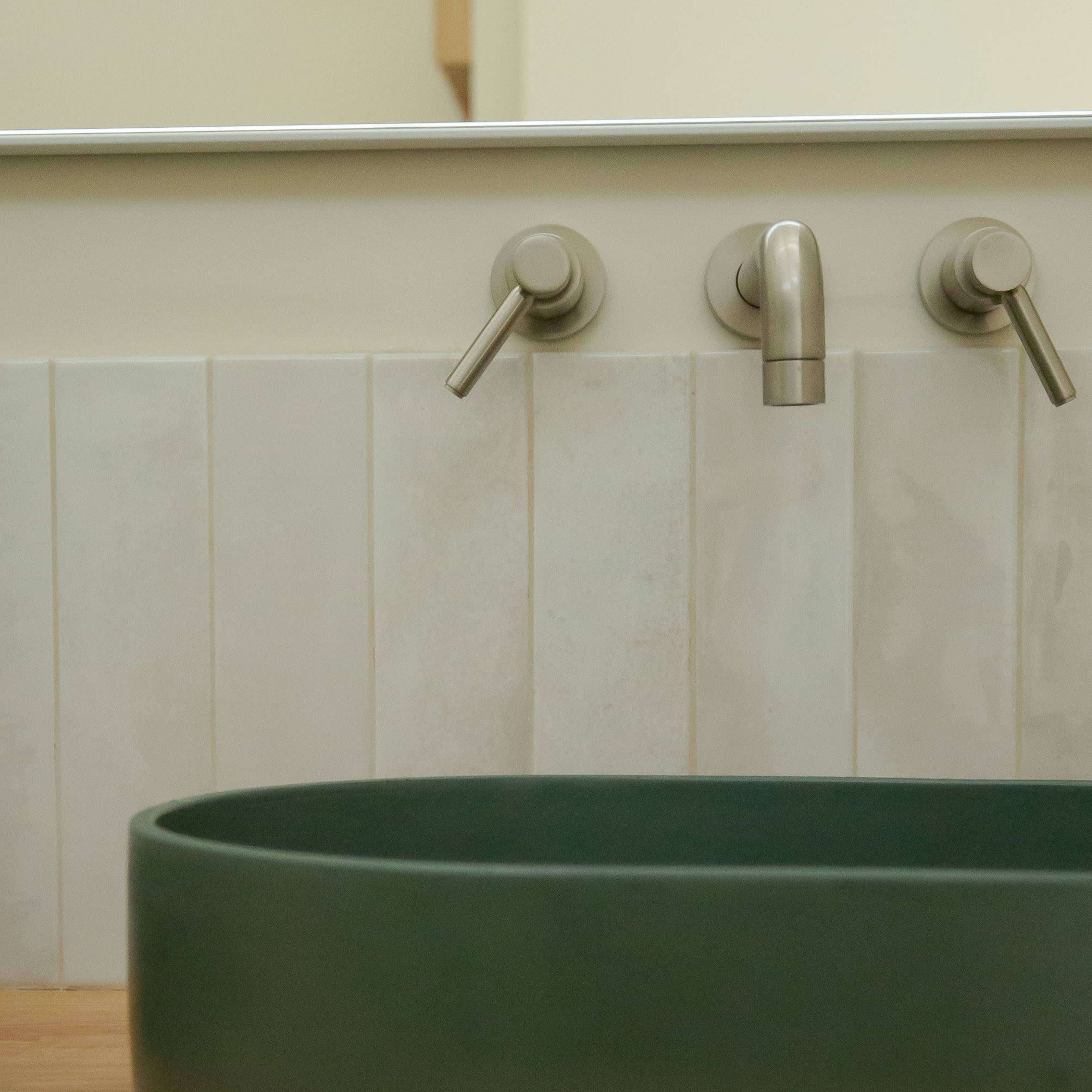 A green oval sink is positioned in front of a wall with vertical white tiles and two metal faucets.