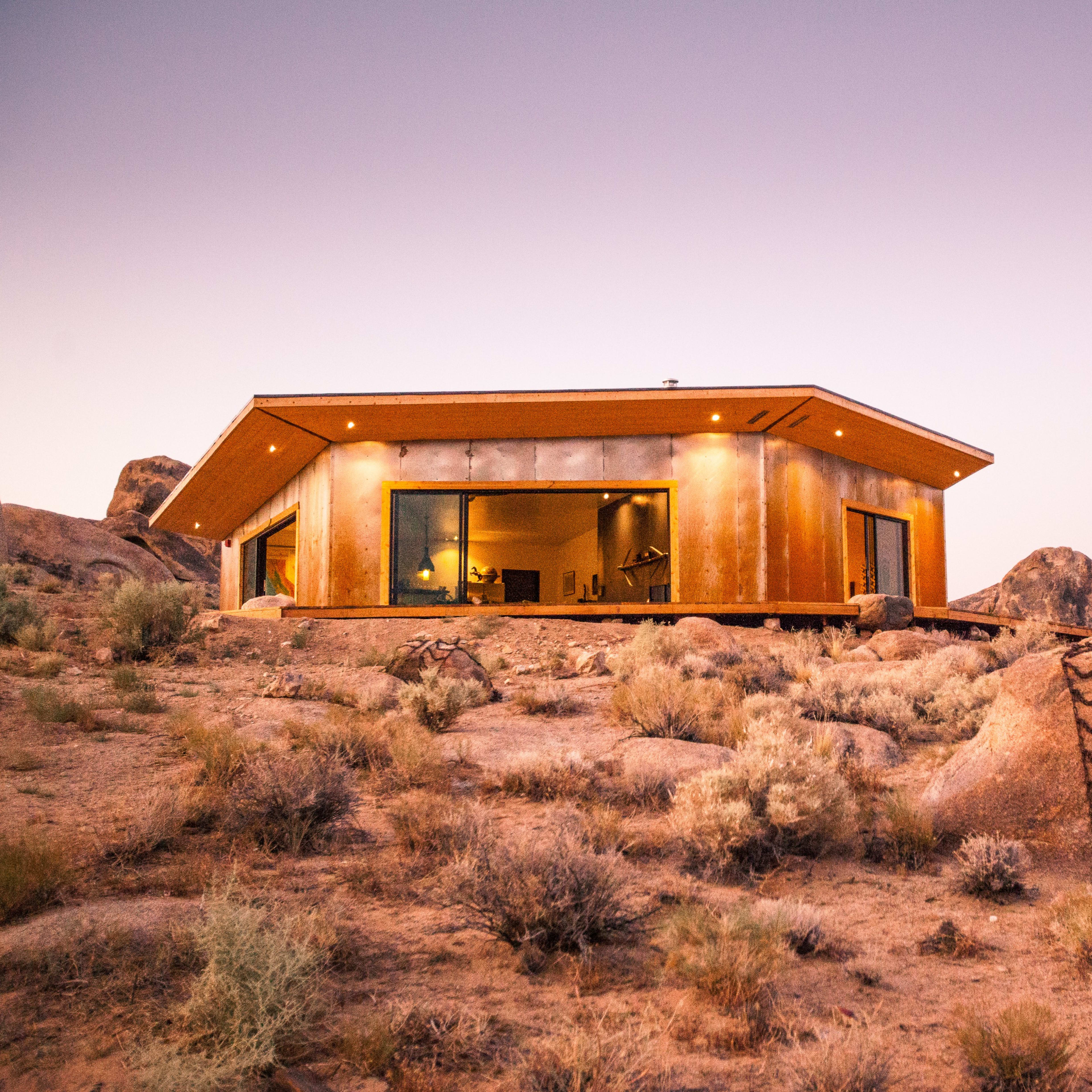 A modern house with large windows sits atop a rocky hill, surrounded by sparse vegetation under a twilight sky.