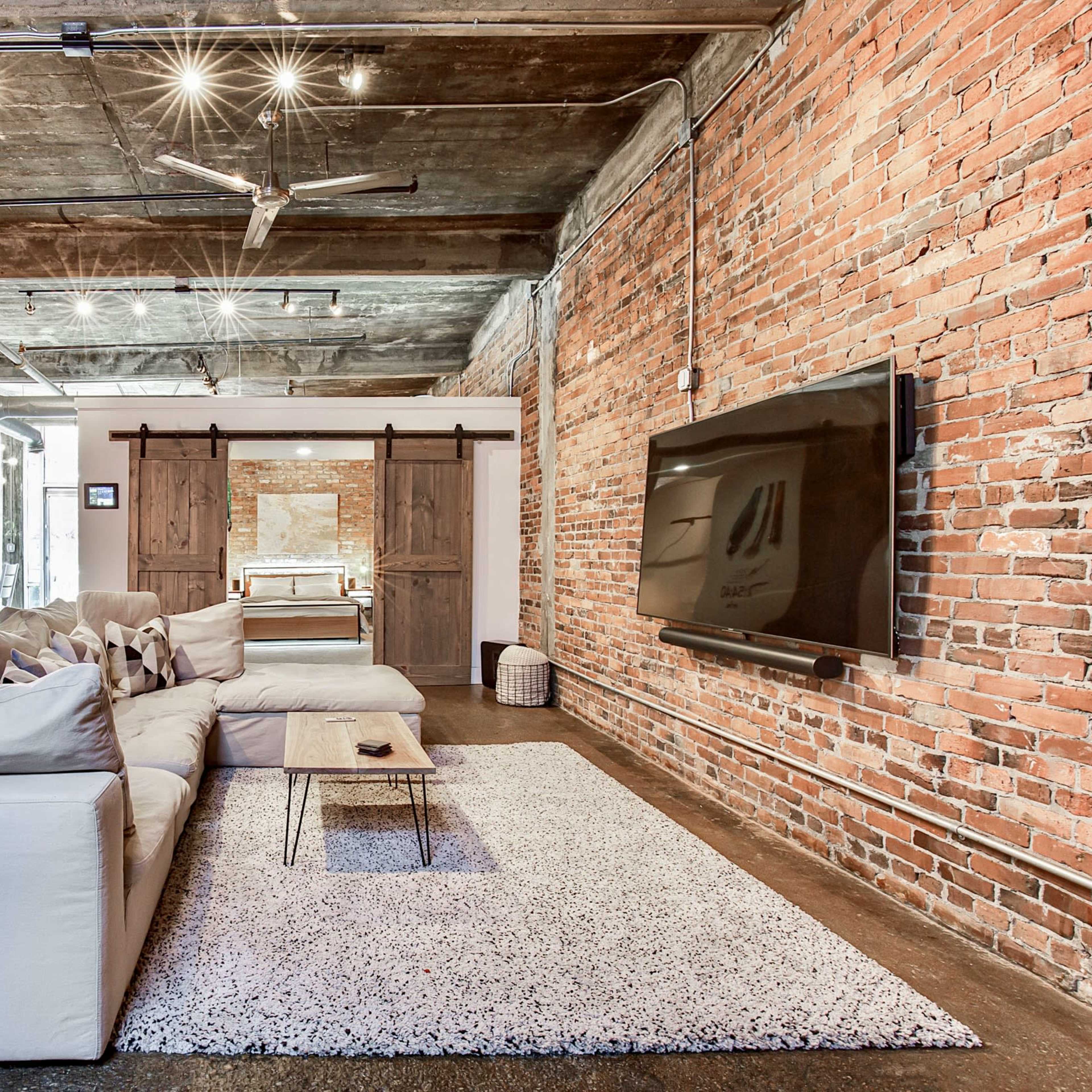 A spacious loft-style living area with exposed brick walls, a large sectional sofa, and a flat-screen TV mounted on the wall, along with a kitchen area visible in the background.