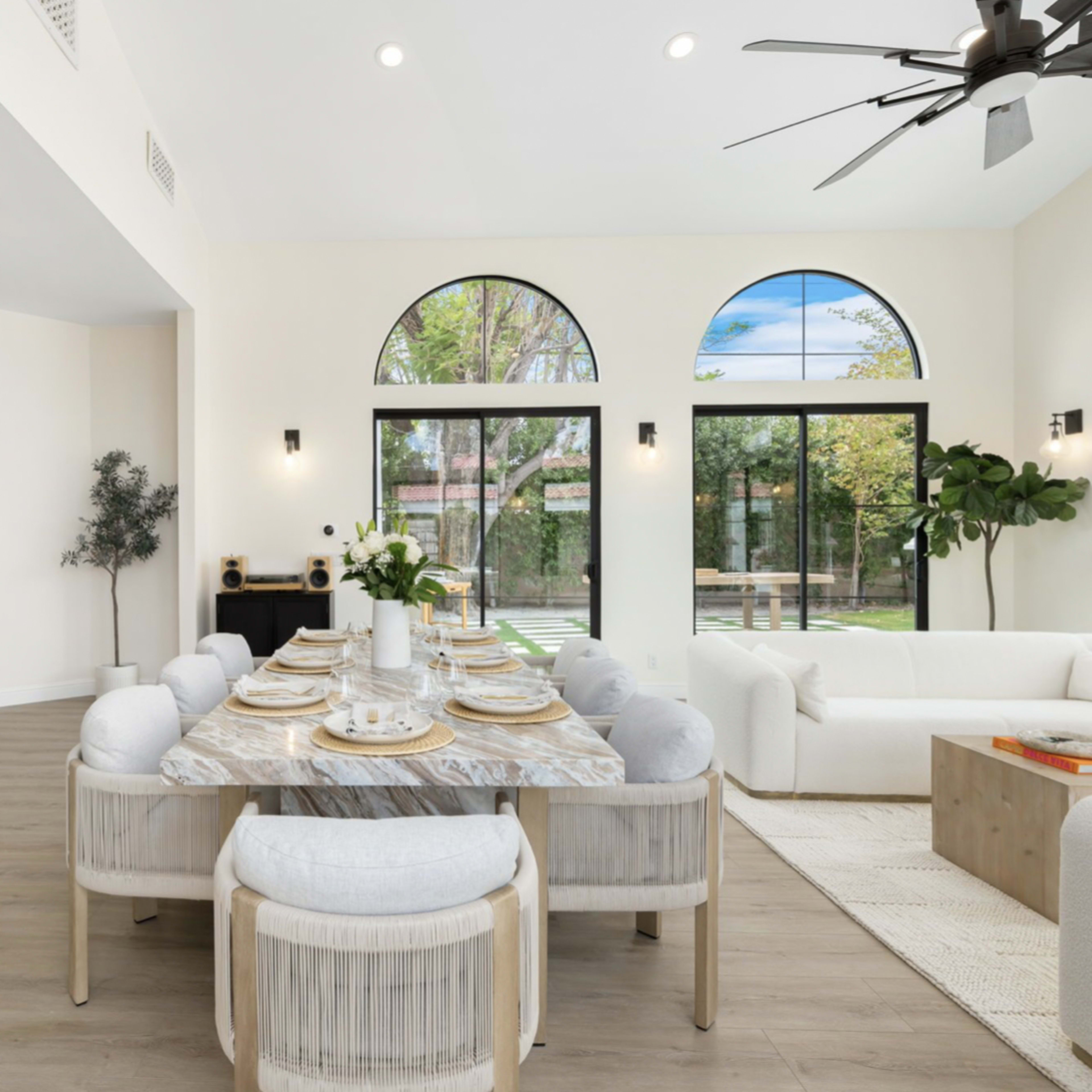 A modern dining area features a round marble table set for six, surrounded by light-colored upholstered chairs, with large windows providing natural light and a view of an outdoor garden.
