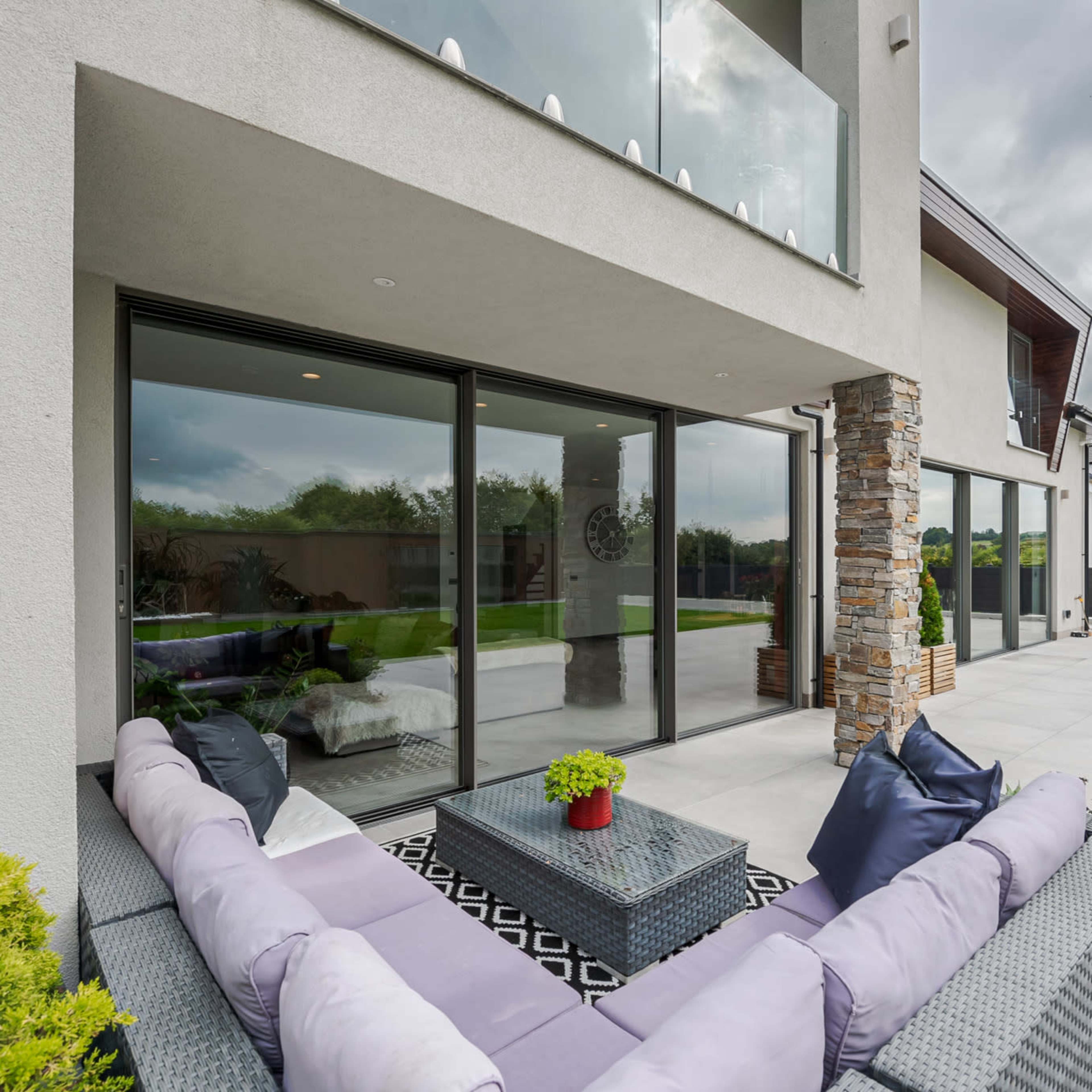 A modern outdoor lounge area with a gray sofa, a glass coffee table, and large sliding glass doors leading into a spacious interior.