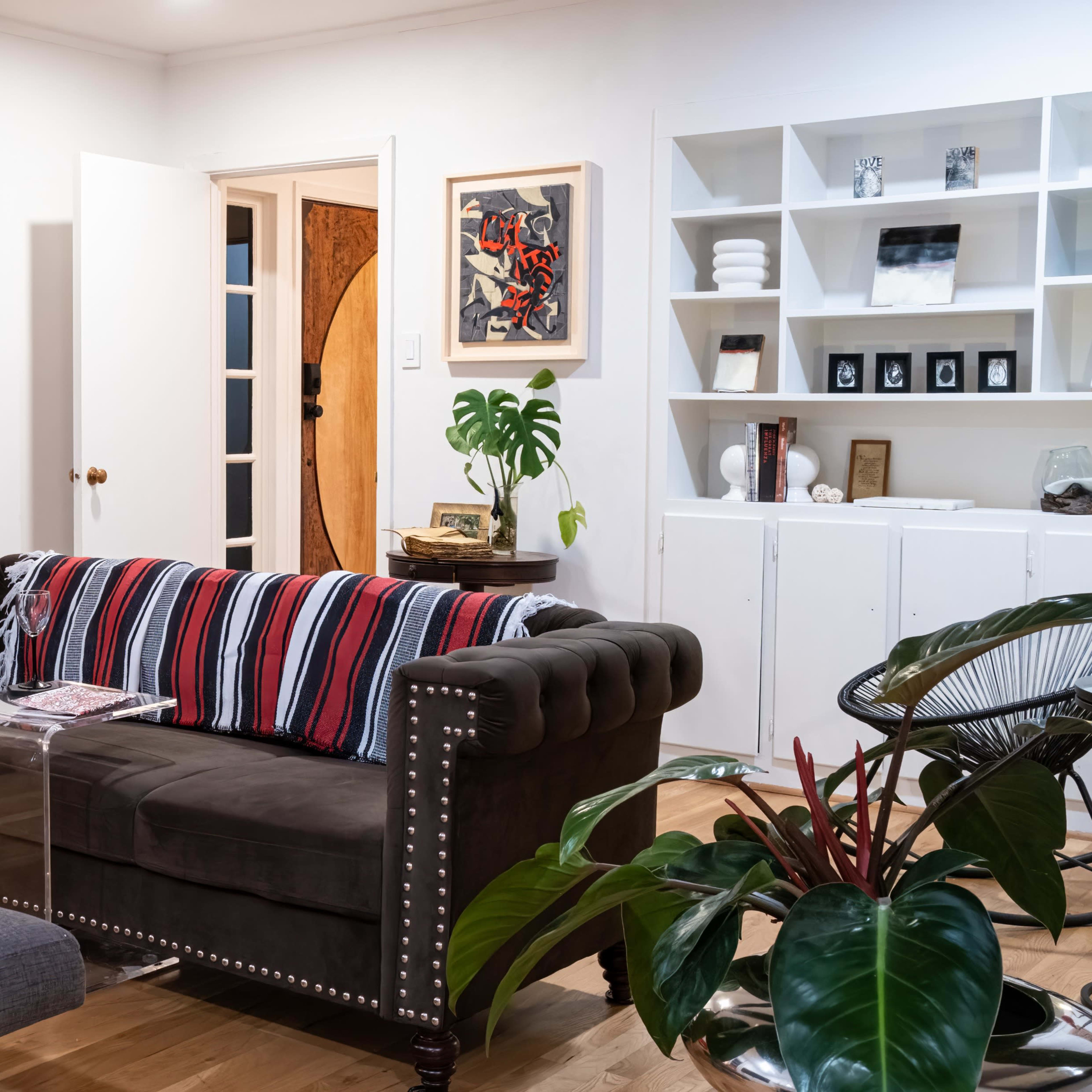 A living room with a tufted brown sofa adorned with striped cushions, a red chair, a glass coffee table, and various artworks on the walls.
