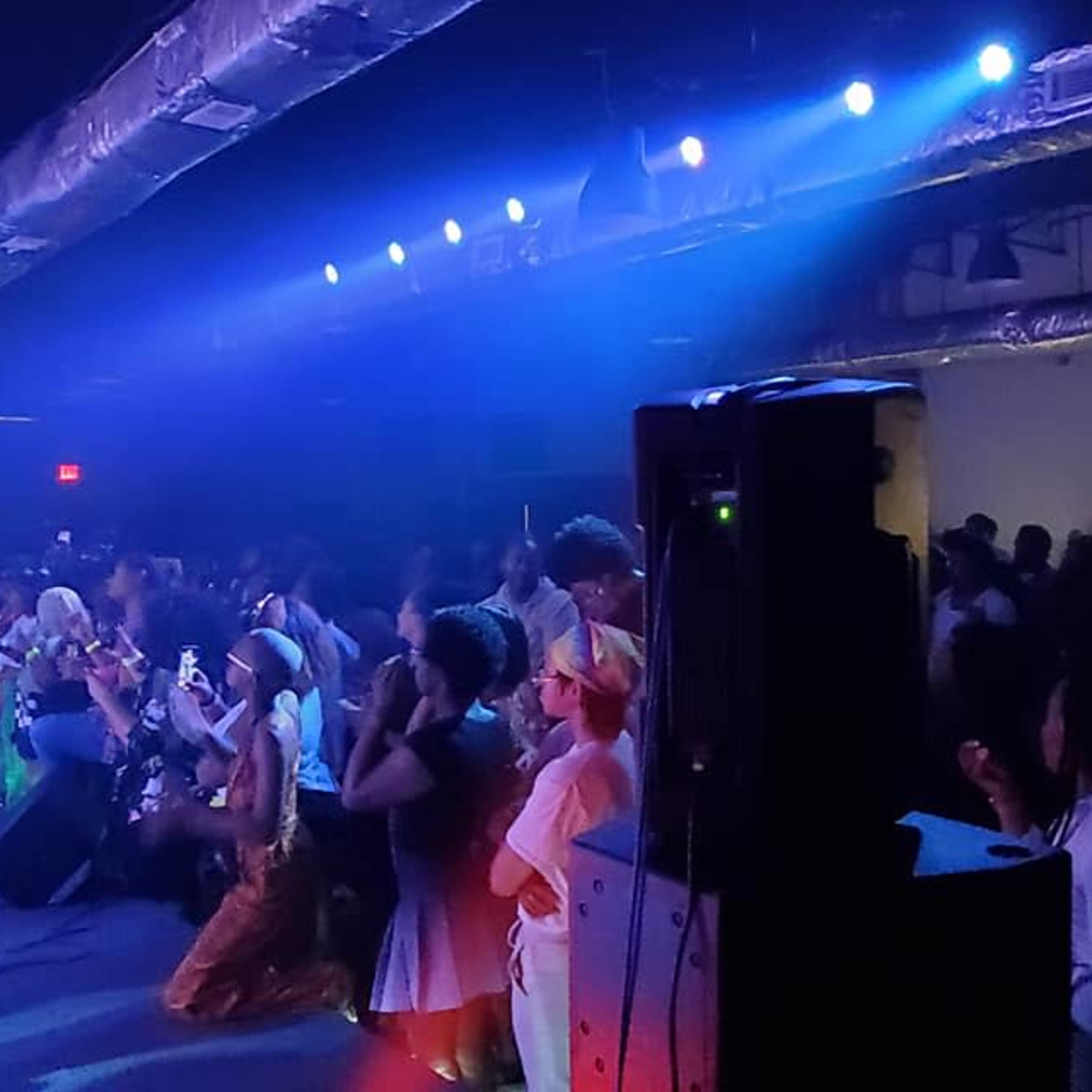 A performer in a hat sings on stage while an audience watches and dances in a dimly lit venue.
