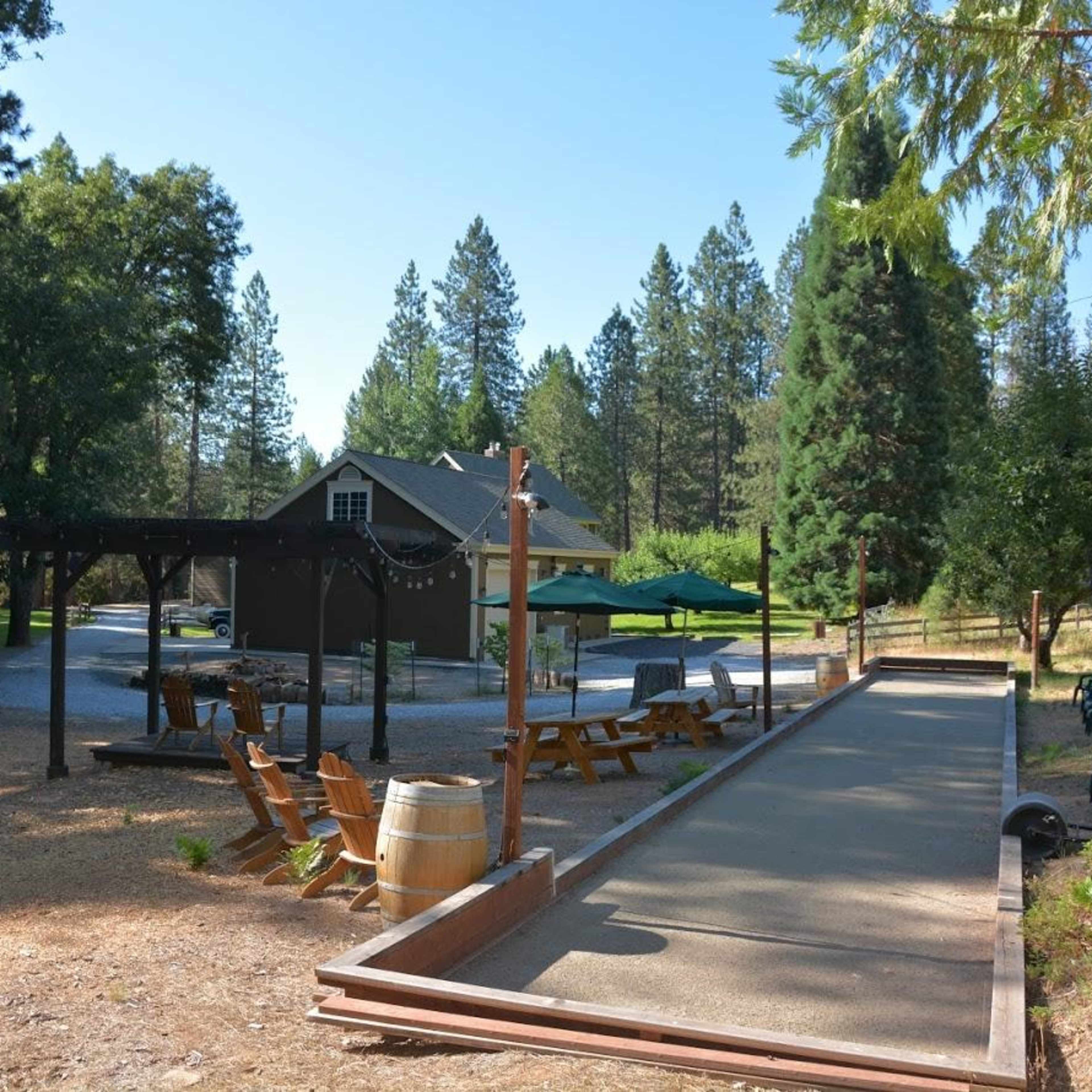 An outdoor recreational area featuring a bocce ball court, wooden seating, and a rustic building surrounded by tall trees.