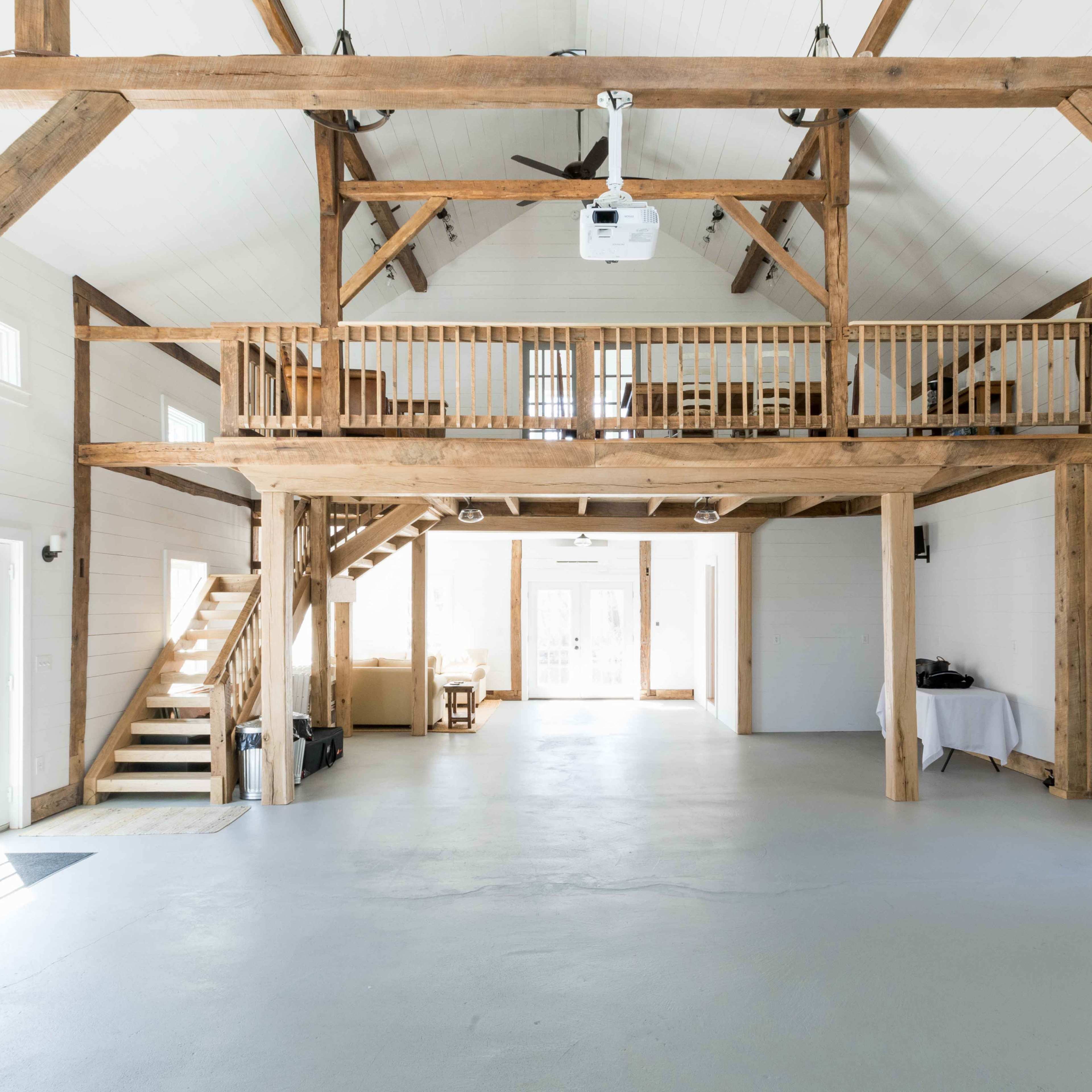 An open interior space featuring exposed wooden beams, a loft area, and large windows that allow natural light to fill the room.