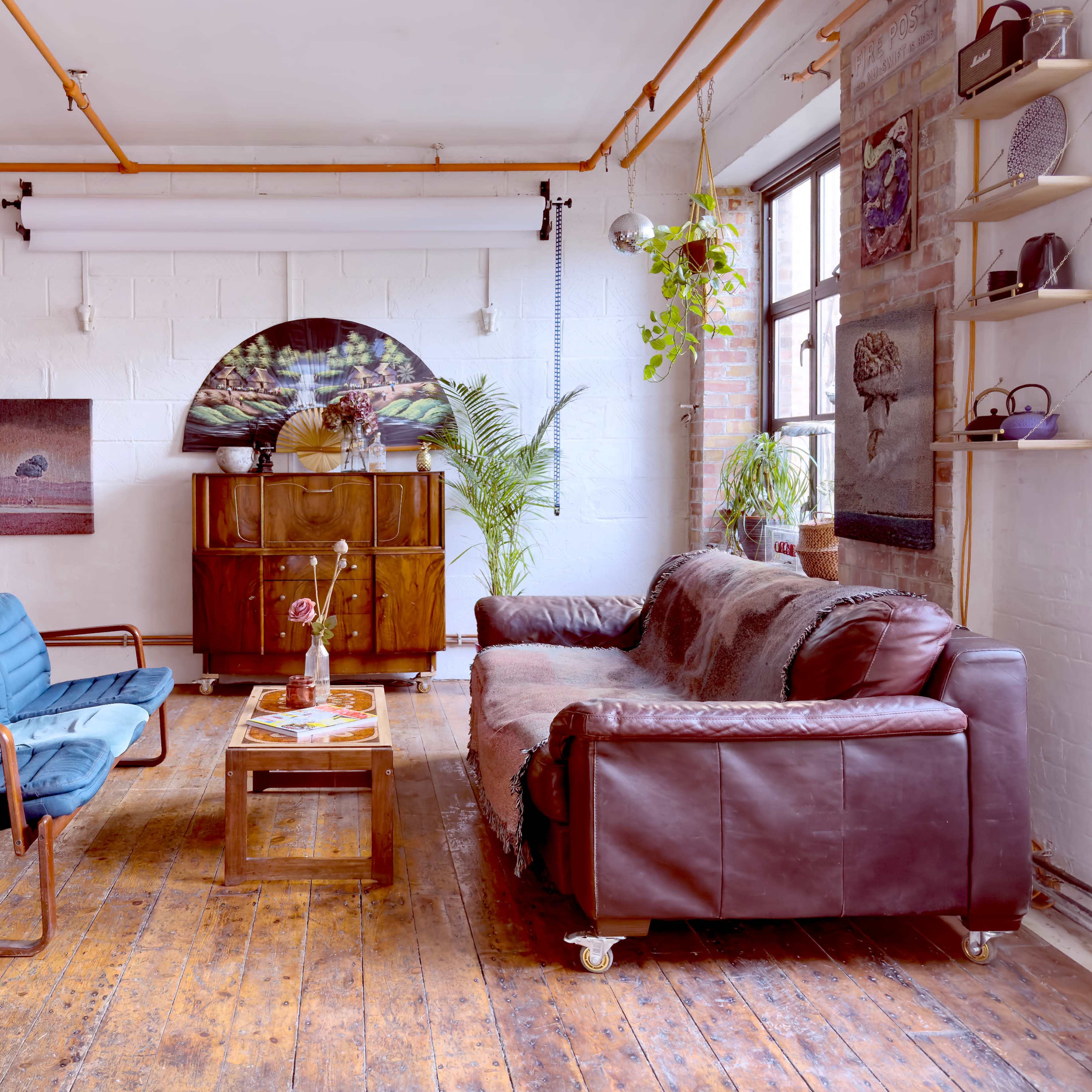 A living room features a blue armchair and a brown leather sofa set against a backdrop of wooden flooring and a large window with plants.