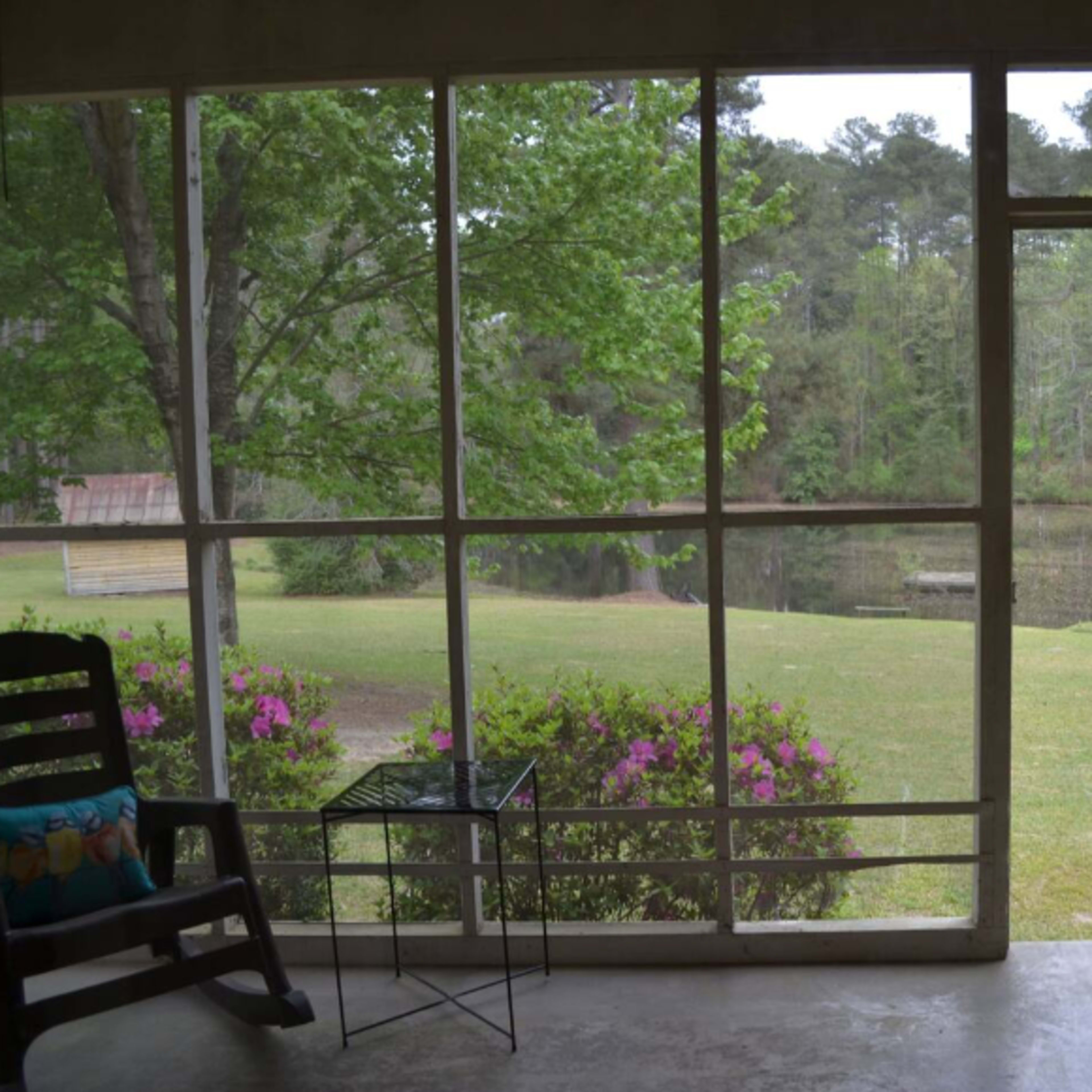 A screened porch with a rocking chair and a small table overlooking a garden and a pond surrounded by trees.