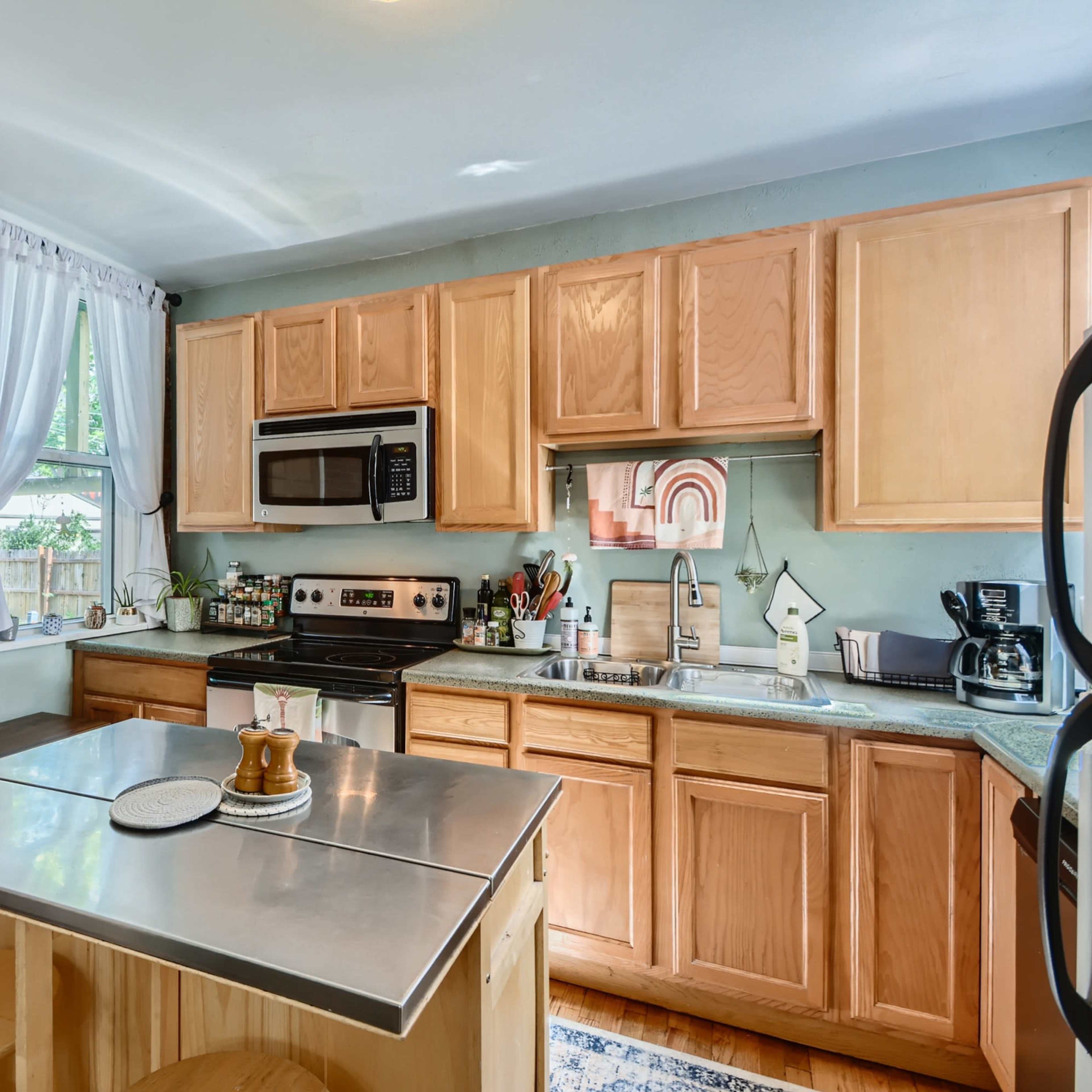 A kitchen features light wood cabinets, stainless steel appliances, a central island with seating, and a window with white curtains allowing natural light.