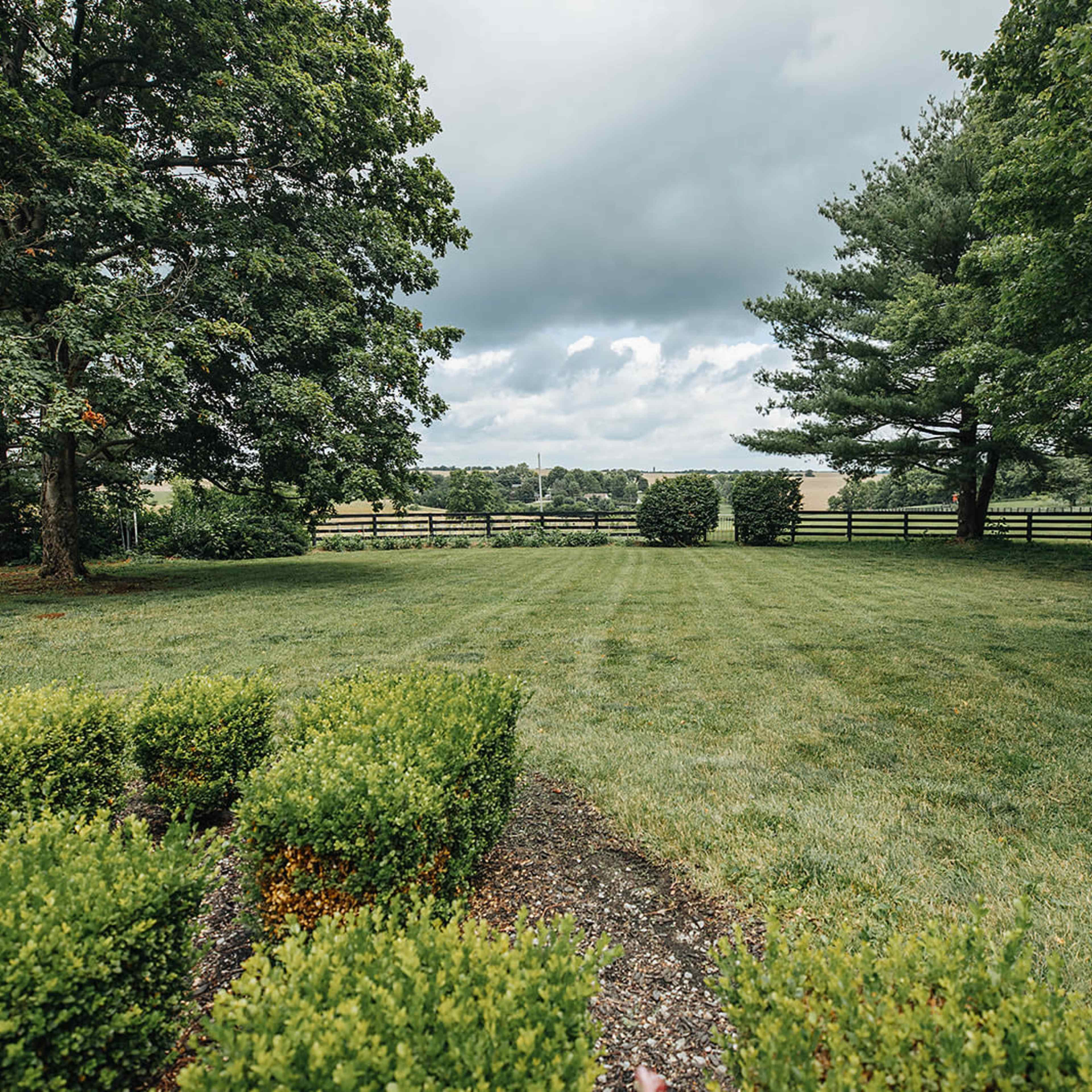 A wide, grassy area is bordered by trees and a wooden fence, with a distant view of hills under a cloudy sky.