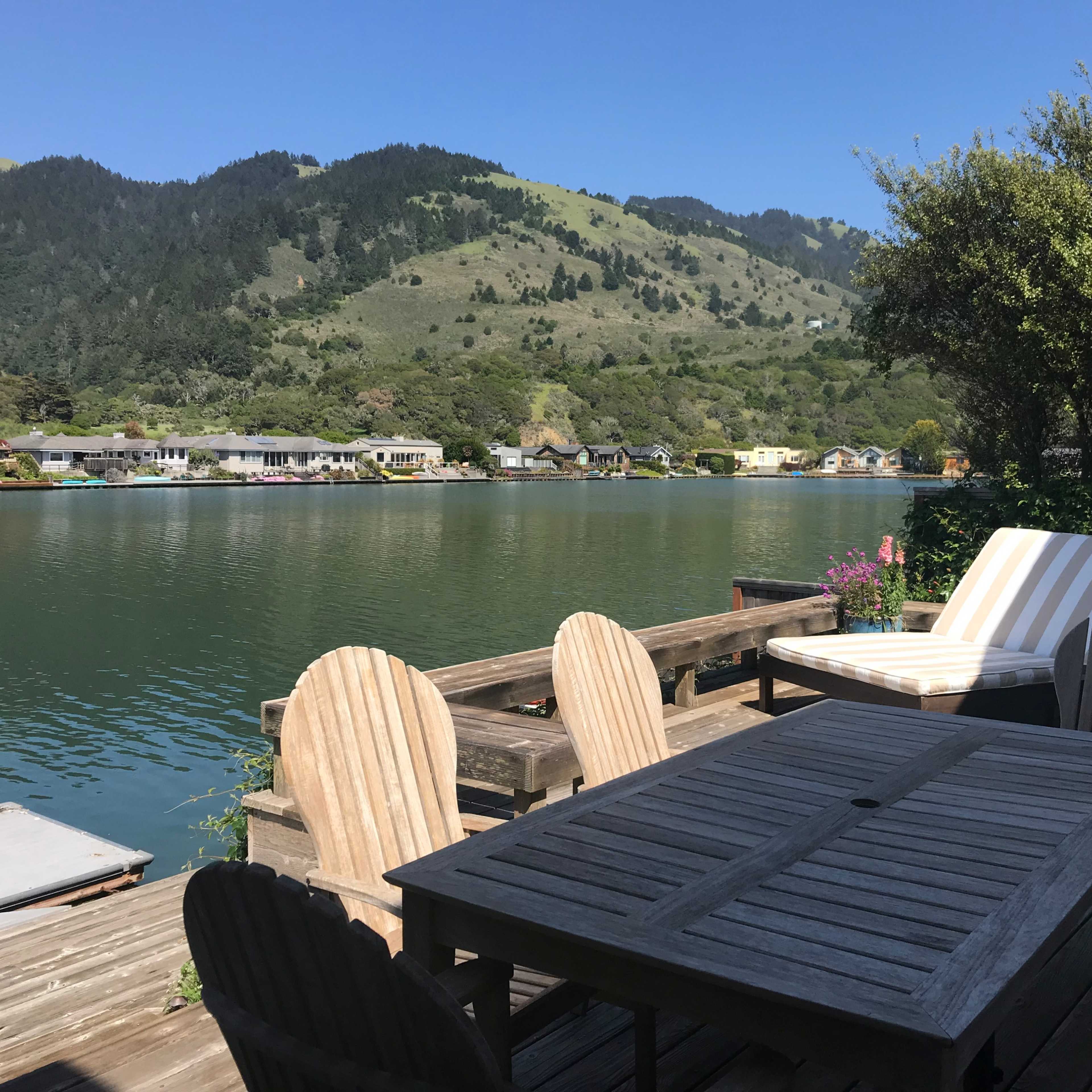 A wooden deck with patio furniture overlooks a calm waterfront and lush green hills in the background.