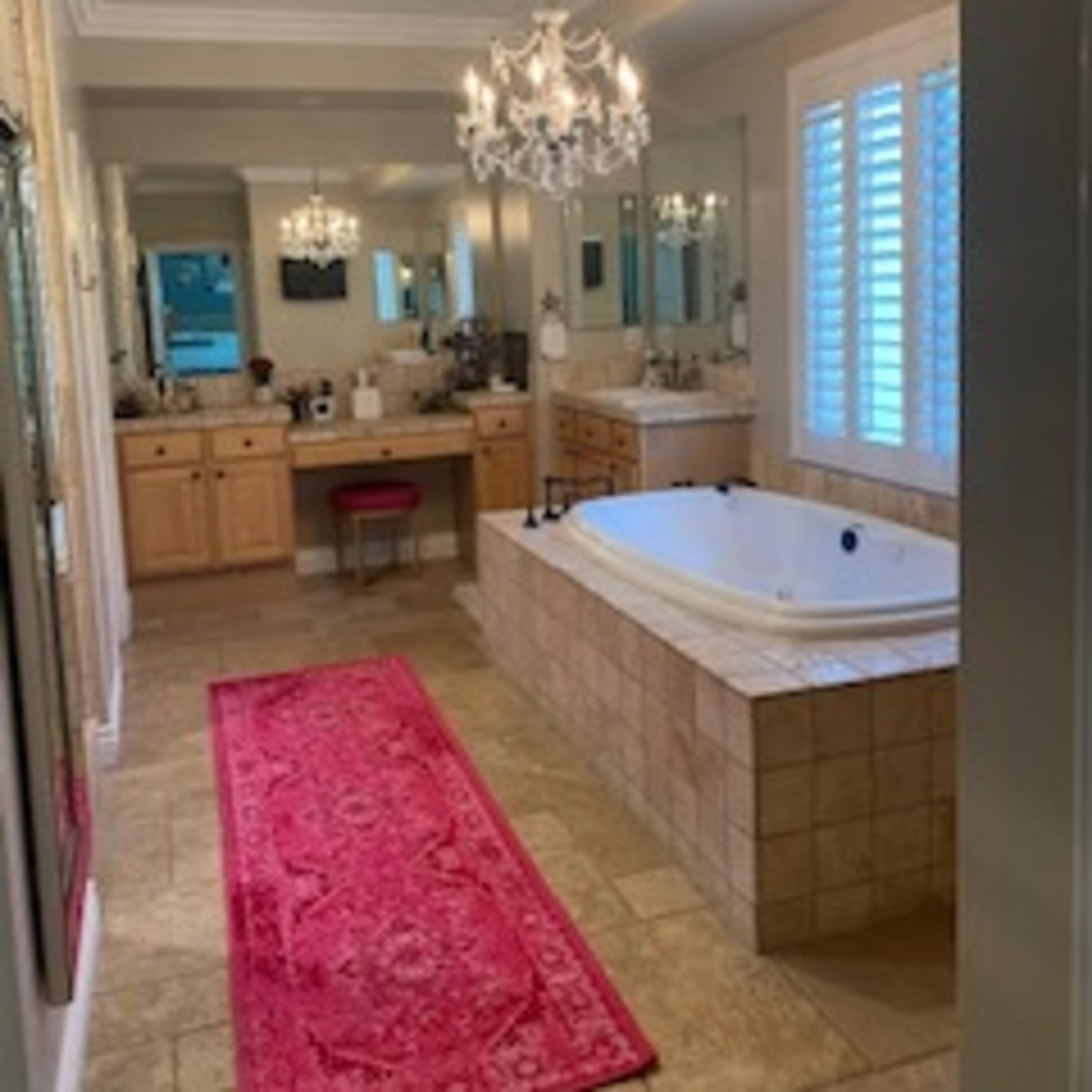 A spacious bathroom featuring a large bathtub, dual sinks, and a decorative chandelier, with a pink runner rug on the tiled floor.