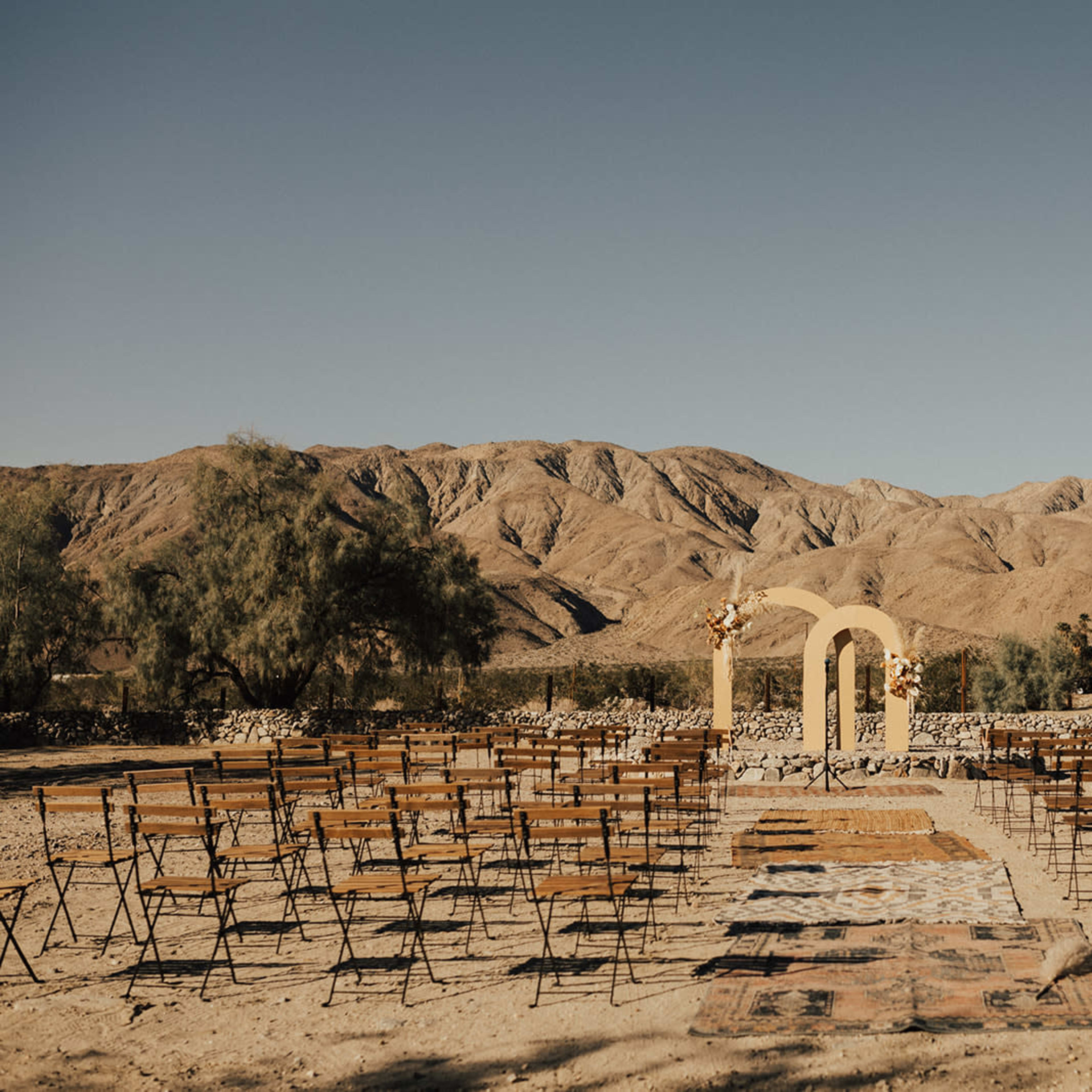 A wedding ceremony setup with rows of wooden chairs facing a decorated arch in a mountainous desert landscape.