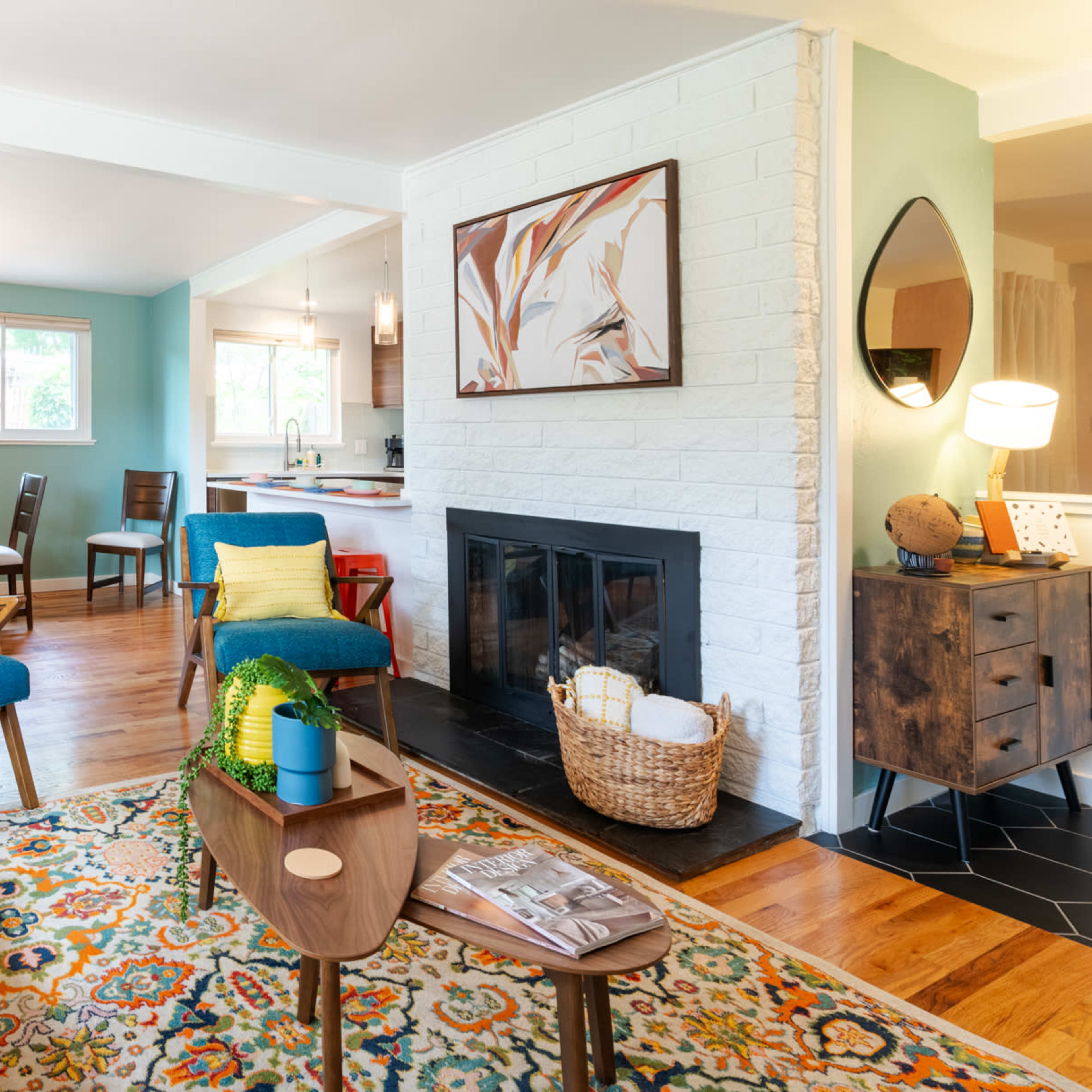 A modern living room features a yellow sofa, blue accent chairs, a patterned rug, and a fireplace, with a dining area visible in the background.