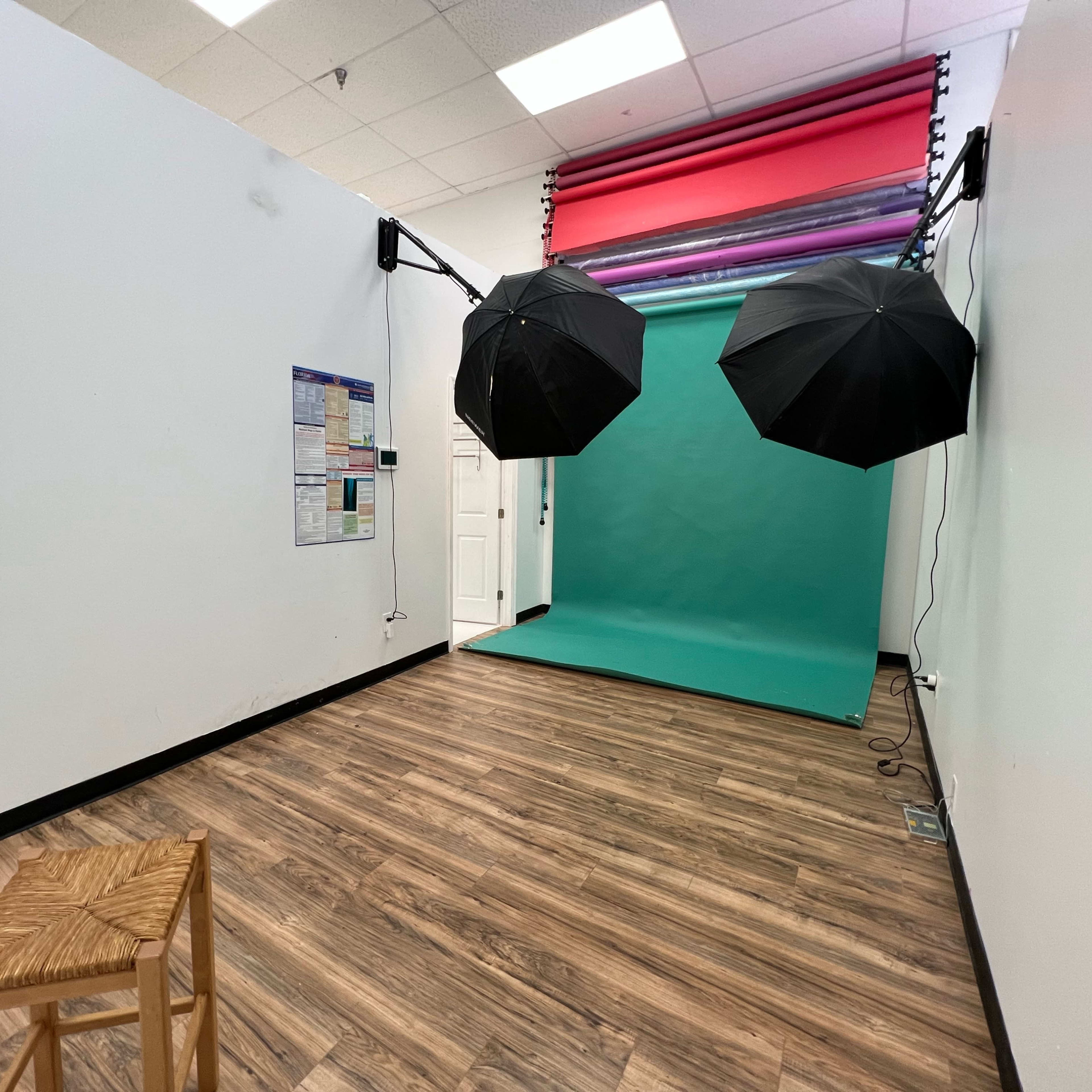 A small photography studio with a teal backdrop, two black umbrellas for lighting, and a wooden stool.