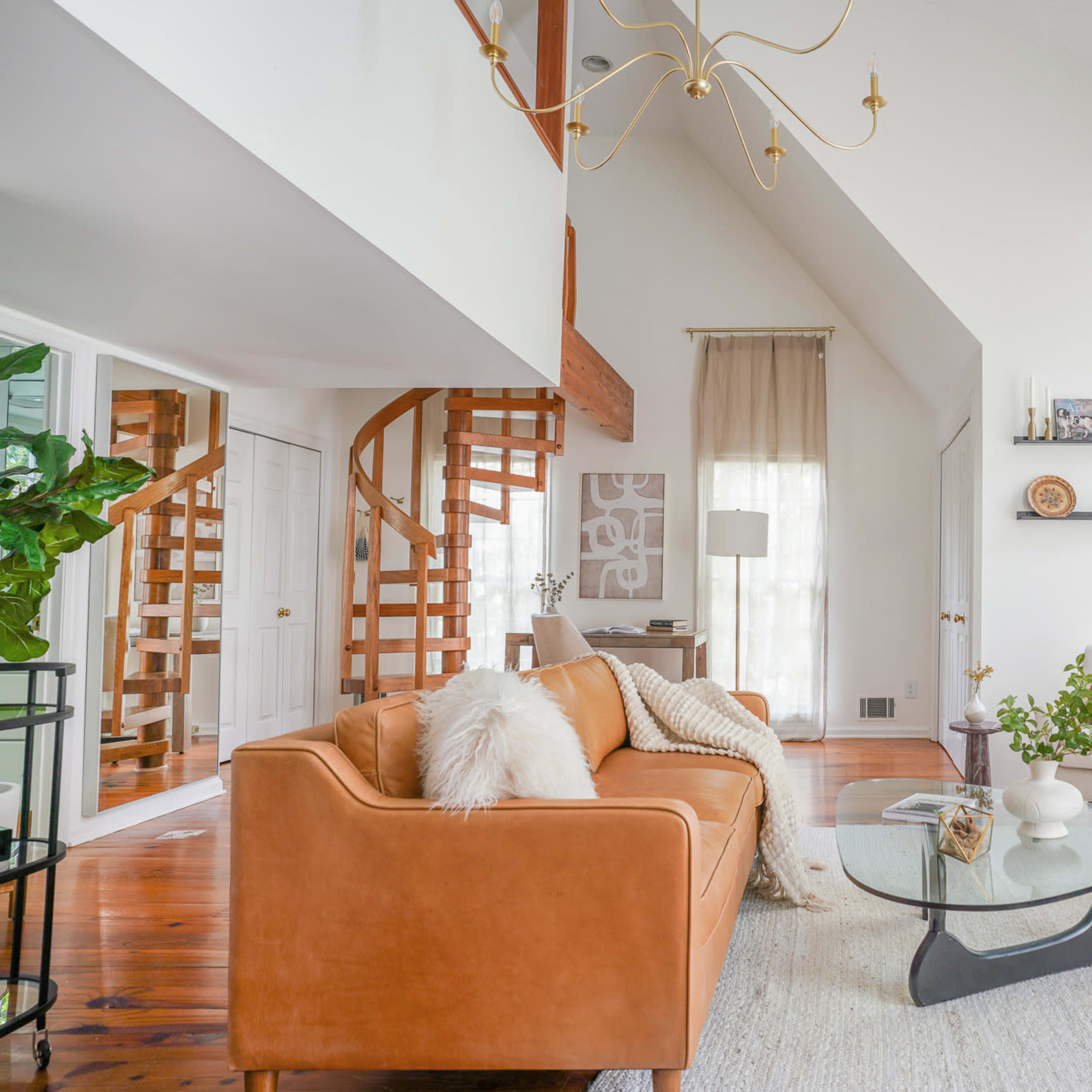 A modern living room with a spiral staircase, a brown leather sofa, a glass coffee table, and large windows allowing natural light to enter.