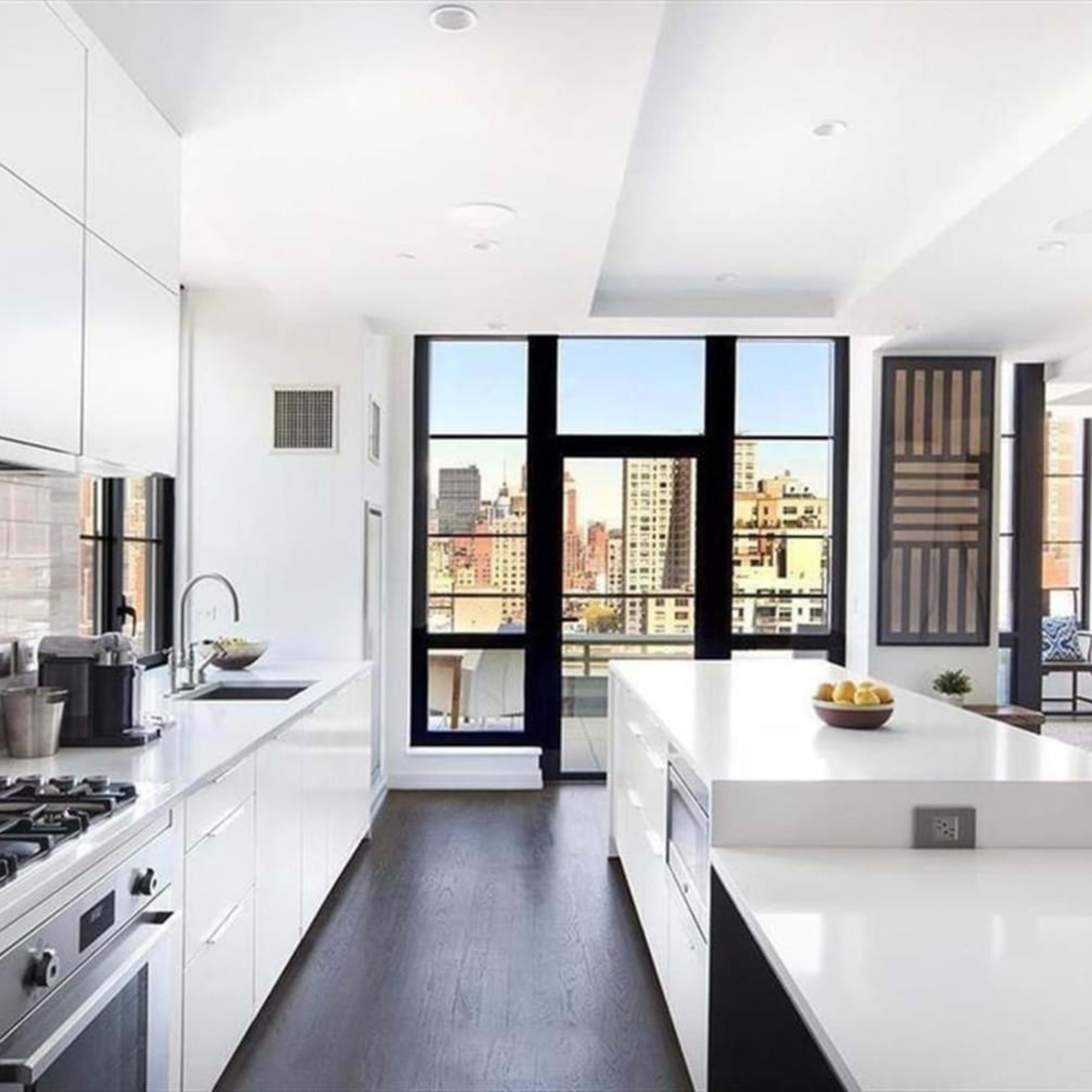 A modern kitchen features sleek white cabinets, a large island with a sink, and floor-to-ceiling windows overlooking a cityscape.