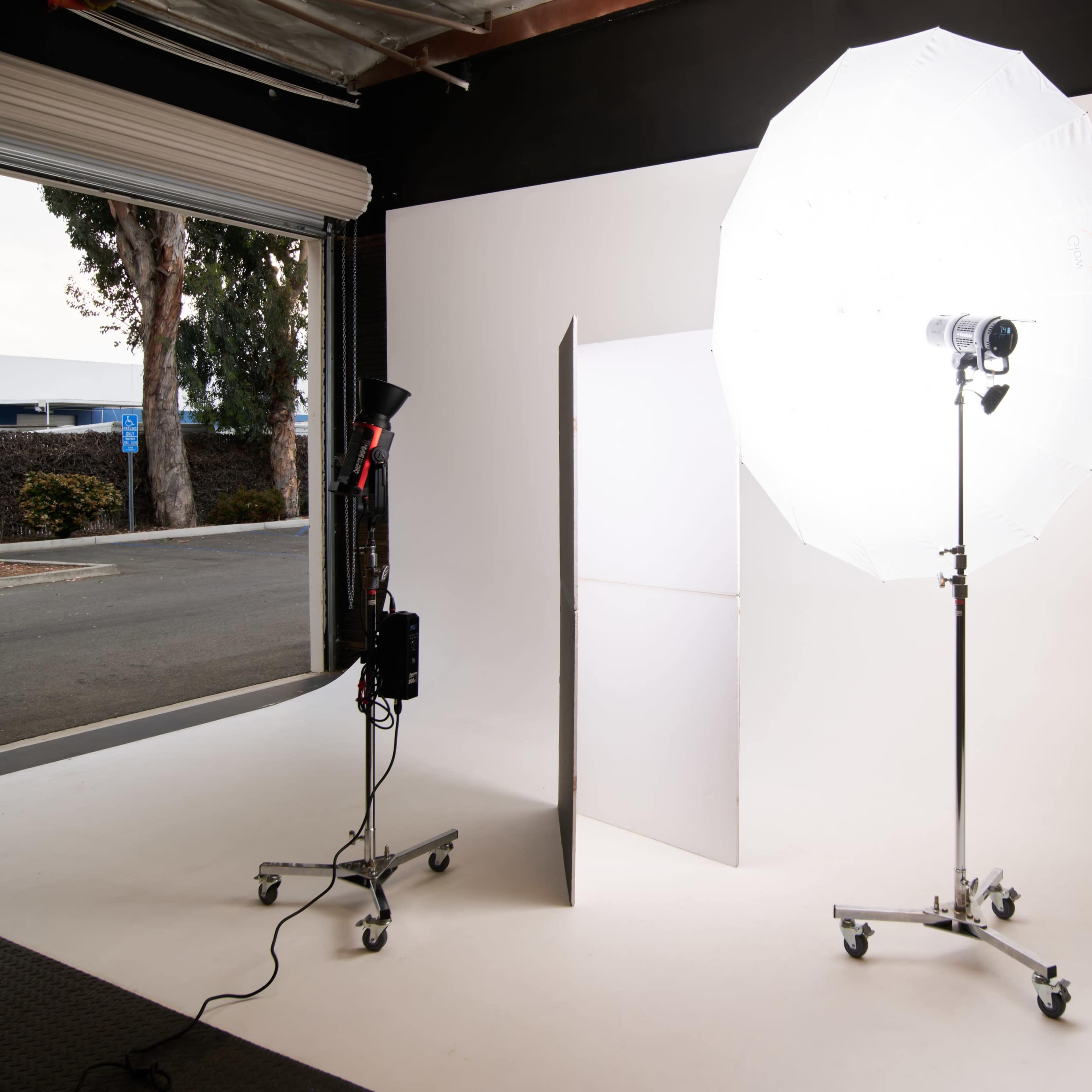 A photography studio setup features a large umbrella light and a reflector stand on a seamless white backdrop.