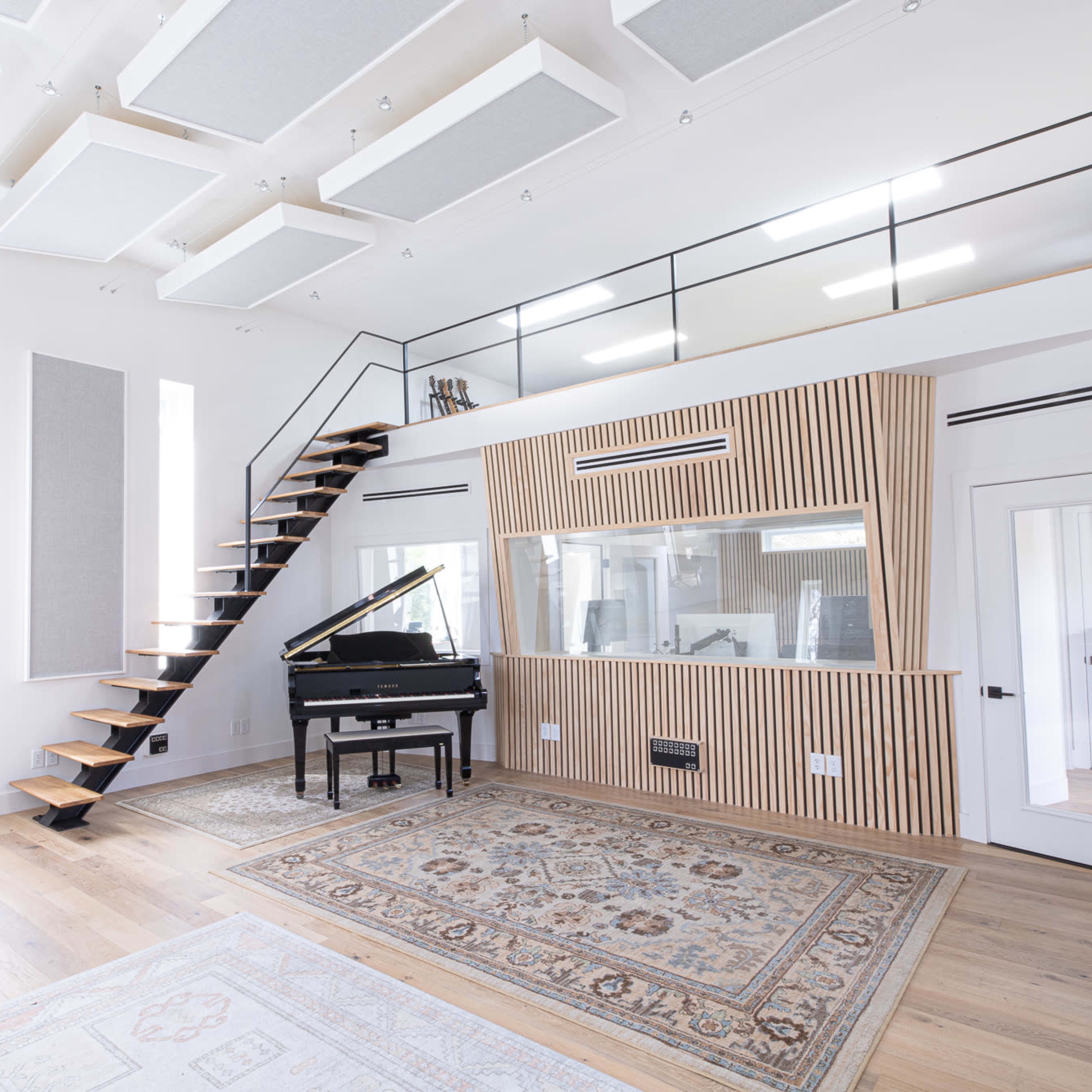 A modern music room featuring a grand piano, a staircase leading to a loft area, and soundproofing panels on the walls.