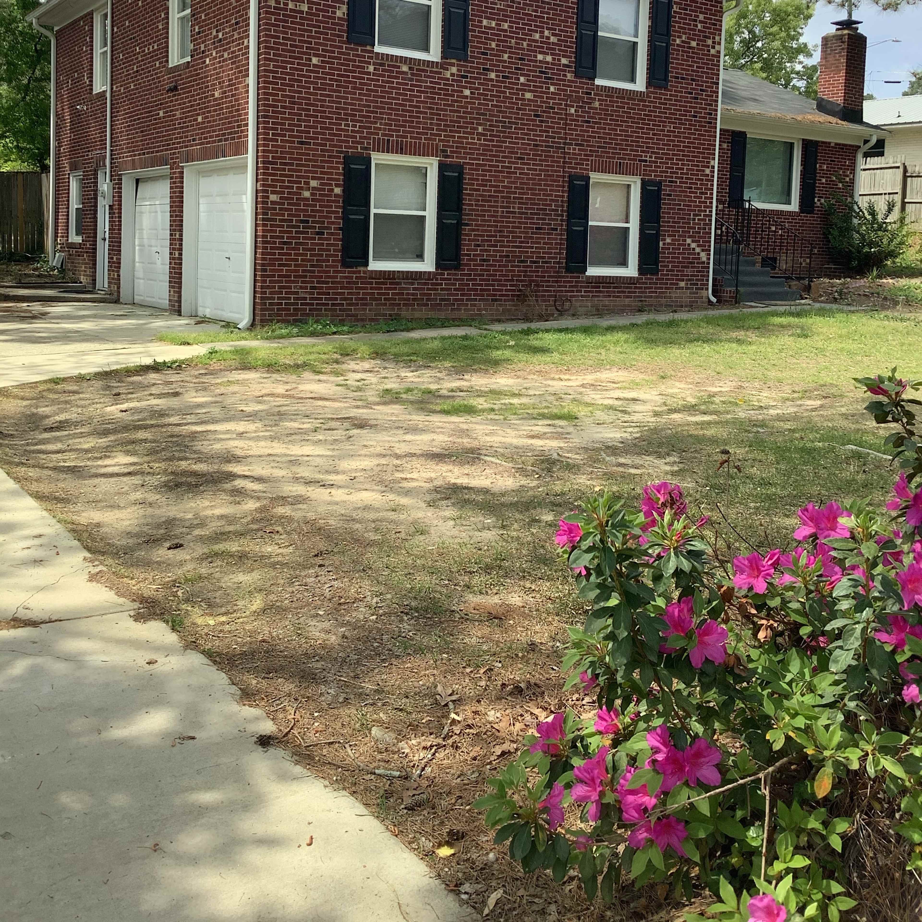 A two-story red brick house with black shutters, a front porch, and a driveway beside flowering pink bushes.
