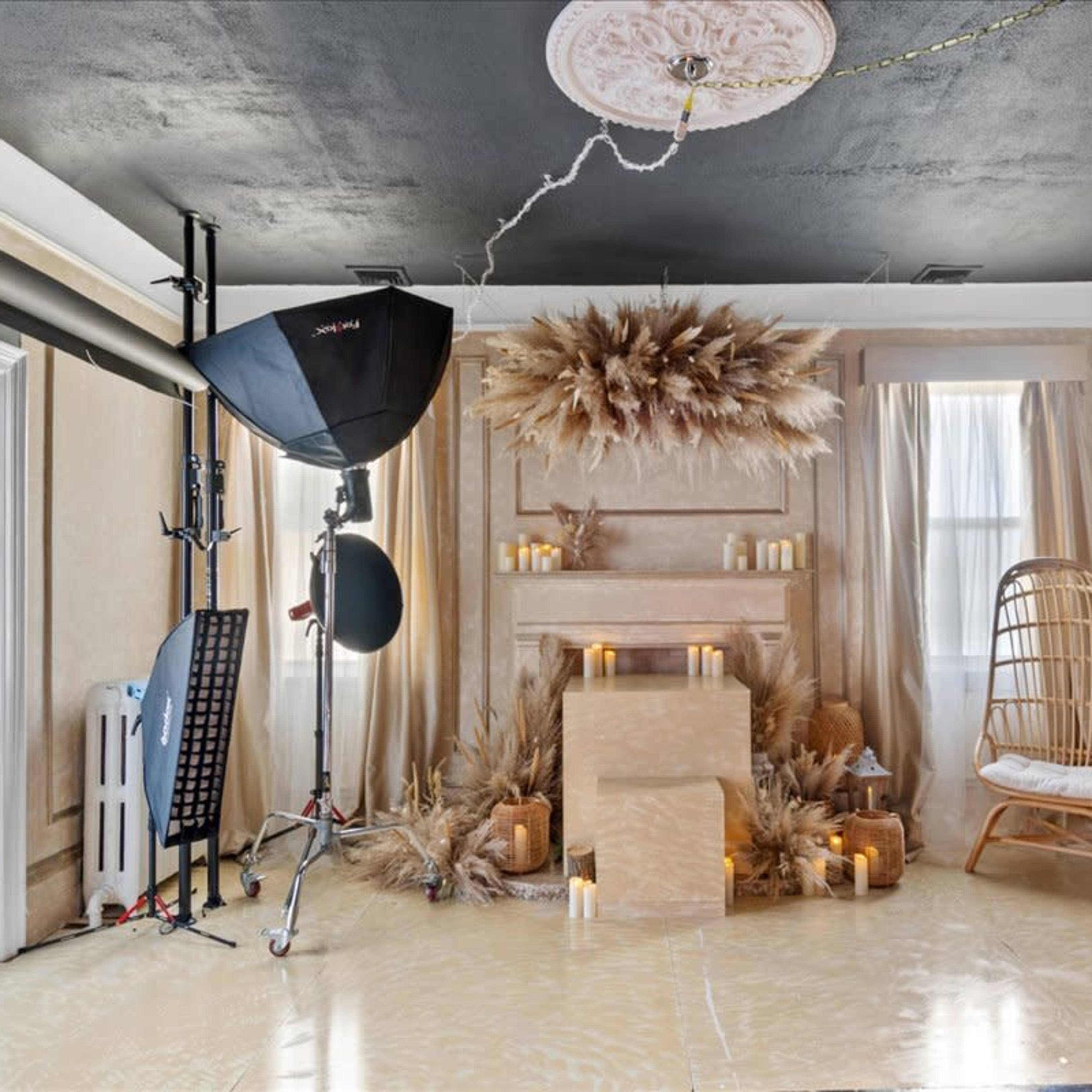 A softly lit studio setup featuring a mantle adorned with candles and decorative elements, a rattan chair, and a large light source on a stand.