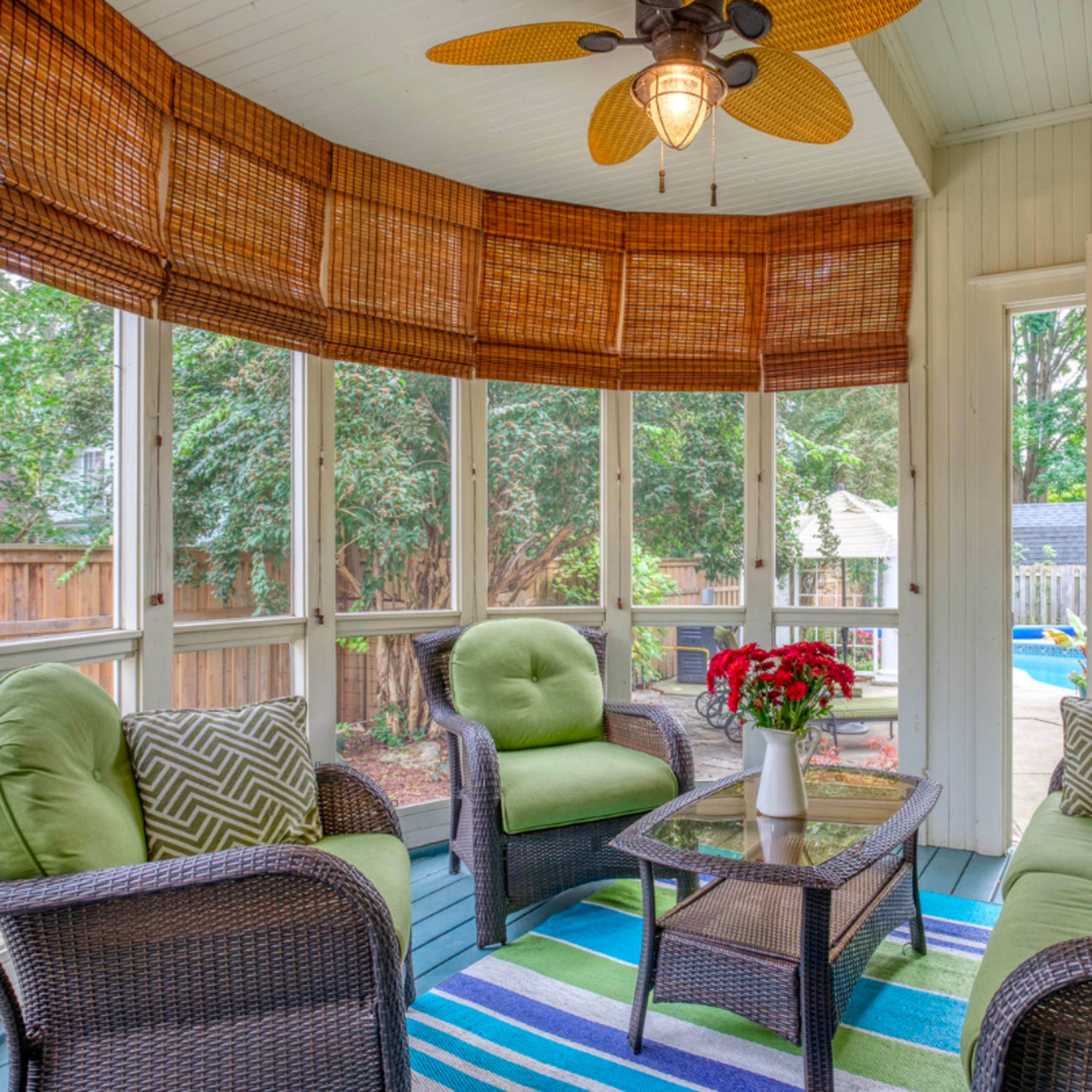 A sunlit screened-in porch furnished with wicker chairs, a small table, a ceiling fan, and a vibrant rug.