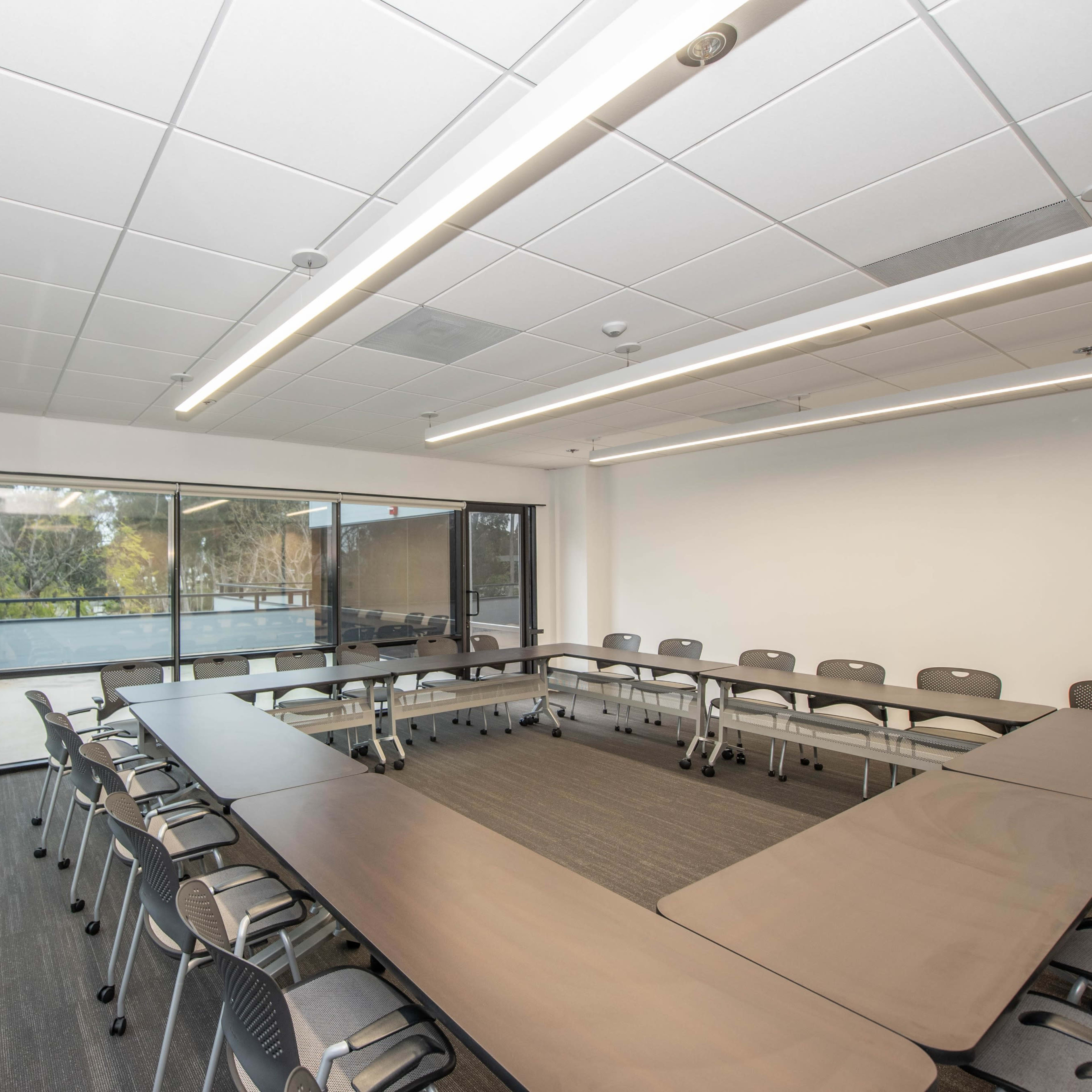 A modern conference room arranged with a large U-shaped table and chairs, illuminated by overhead lights and featuring large windows overlooking greenery.